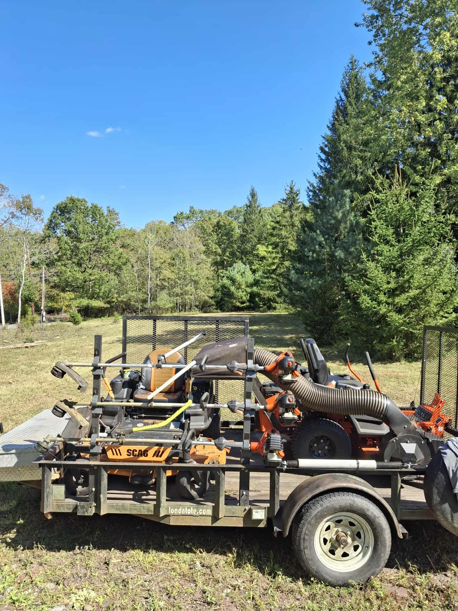 A trailer loaded with landscaping equipment, including a weed trimmer, leaf blower, and leaf vacuum, parked on grass with trees and a blue sky in the background.