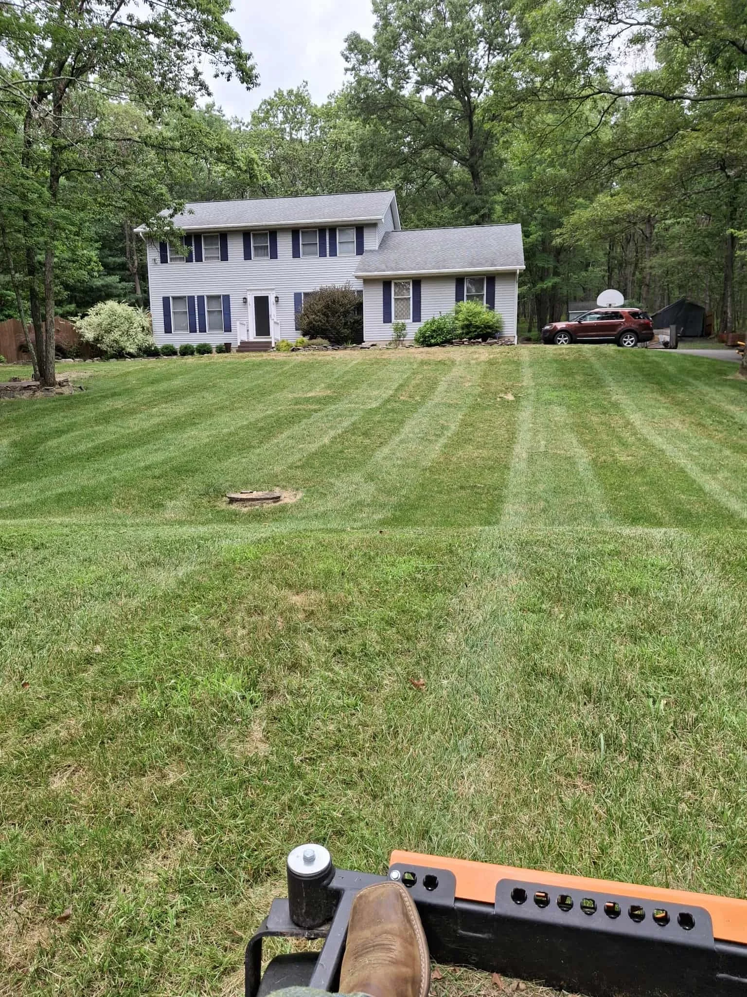 View from a riding lawn mower showing freshly mowed grass leading up to a suburban house, with a car parked in the driveway and trees in the background.