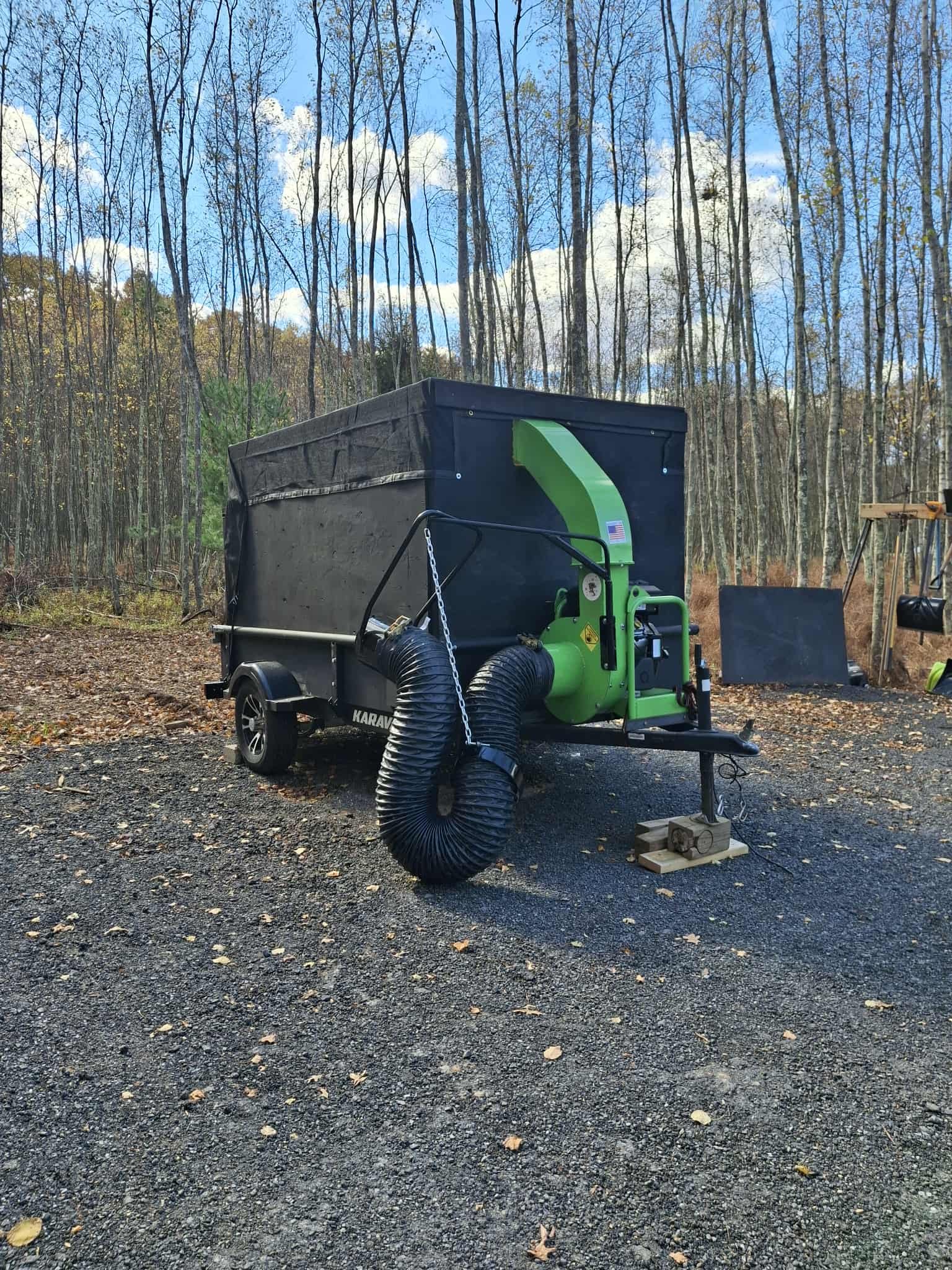 Portable wood chipper on a trailer in a wooded area with autumn foliage, gravel ground, and tools in the background.