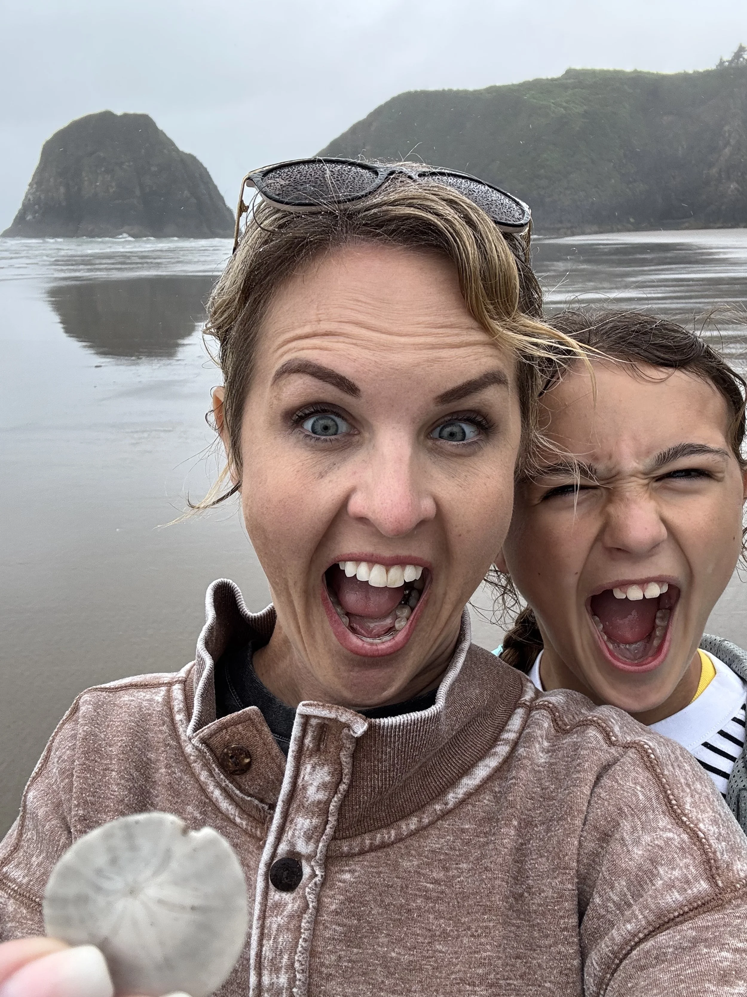 A woman and a young girl on a beach, taking a selfie with the ocean and large rocks in the background. The woman is holding a seashell, both are making surprised or excited facial expressions.