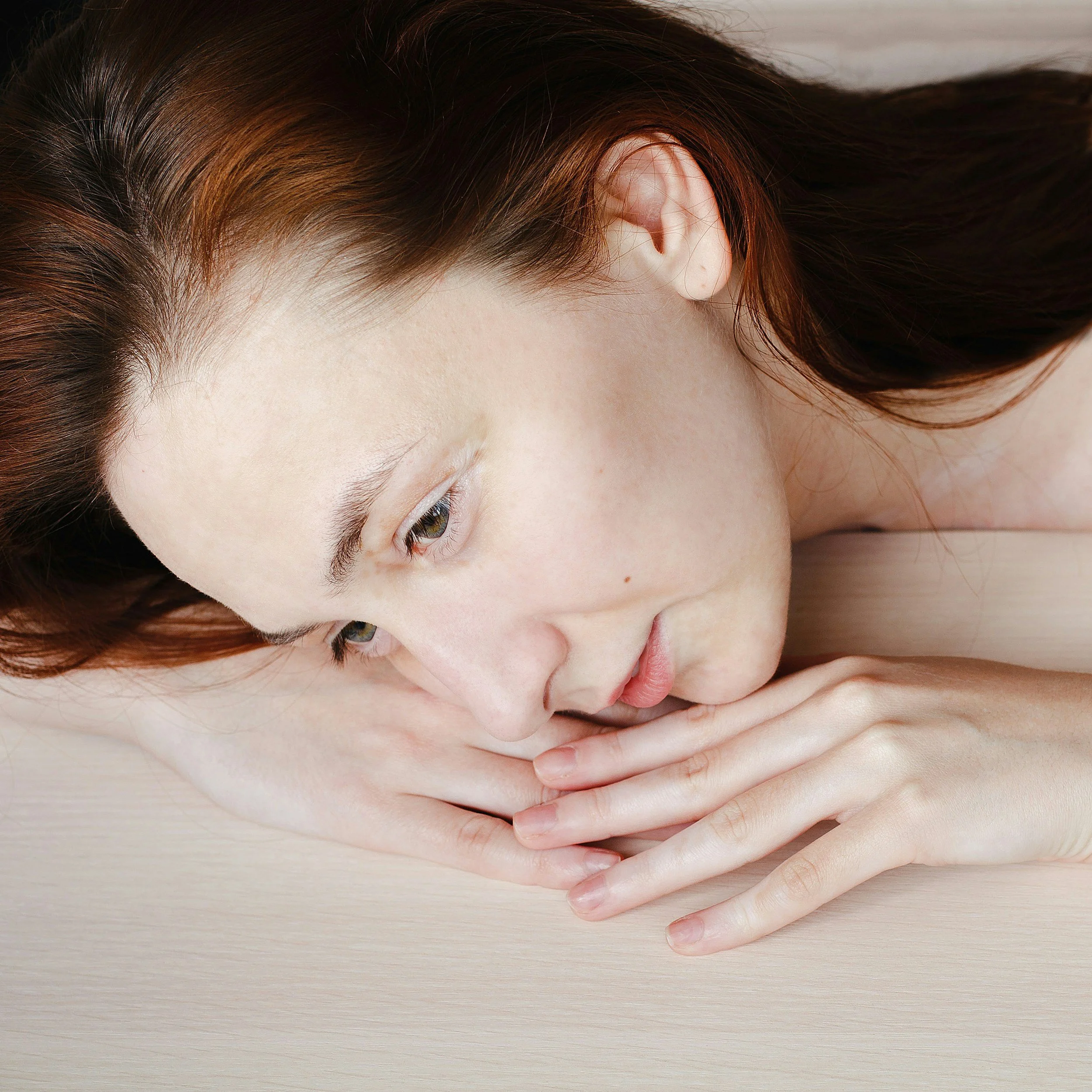 A woman with red hair lying on her side with her head resting on her hands on a light wooden surface.
