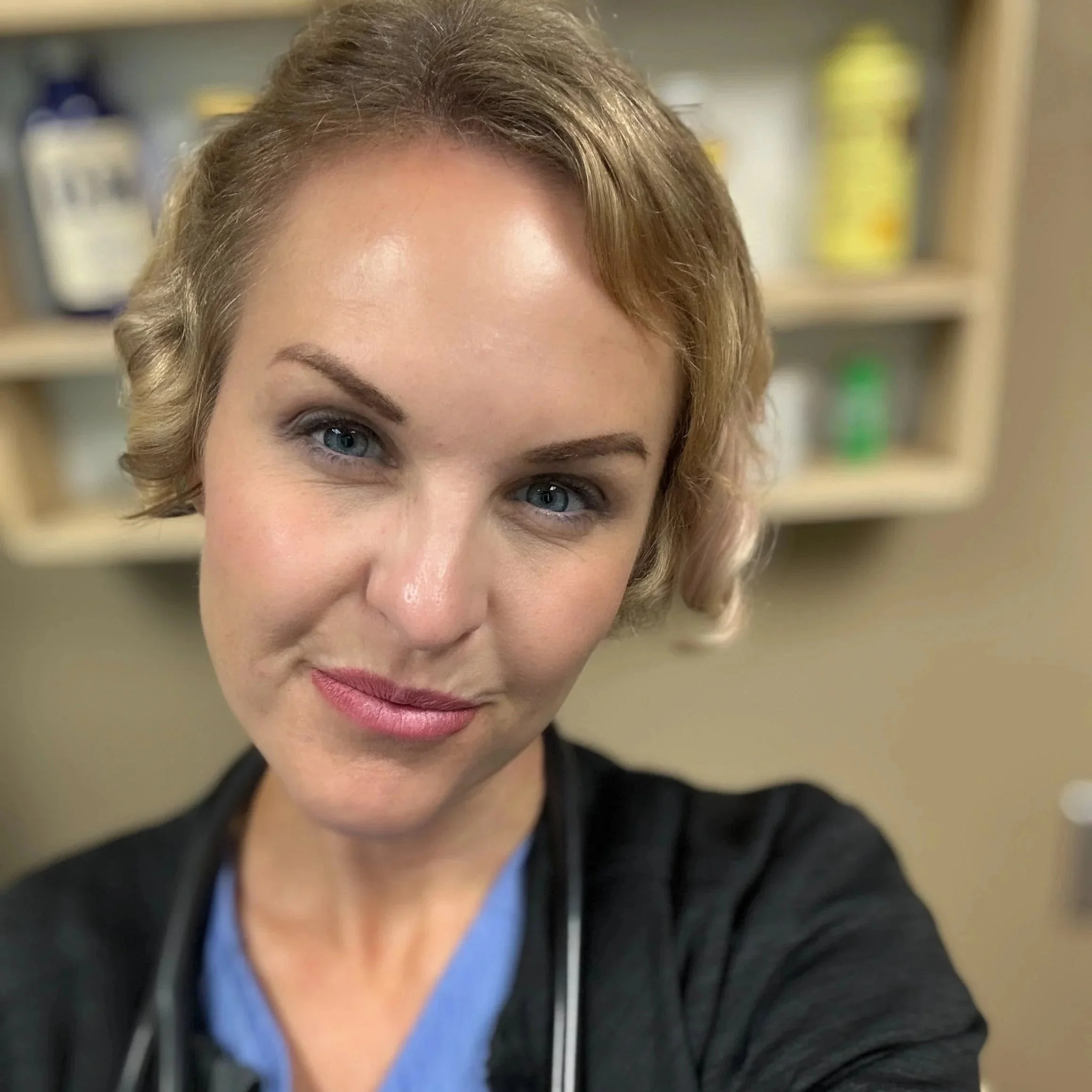 Close-up of a woman with short blonde hair, blue eyes, wearing a blue top and a black jacket, smiling in a medical setting with shelves and medication bottles in the background.