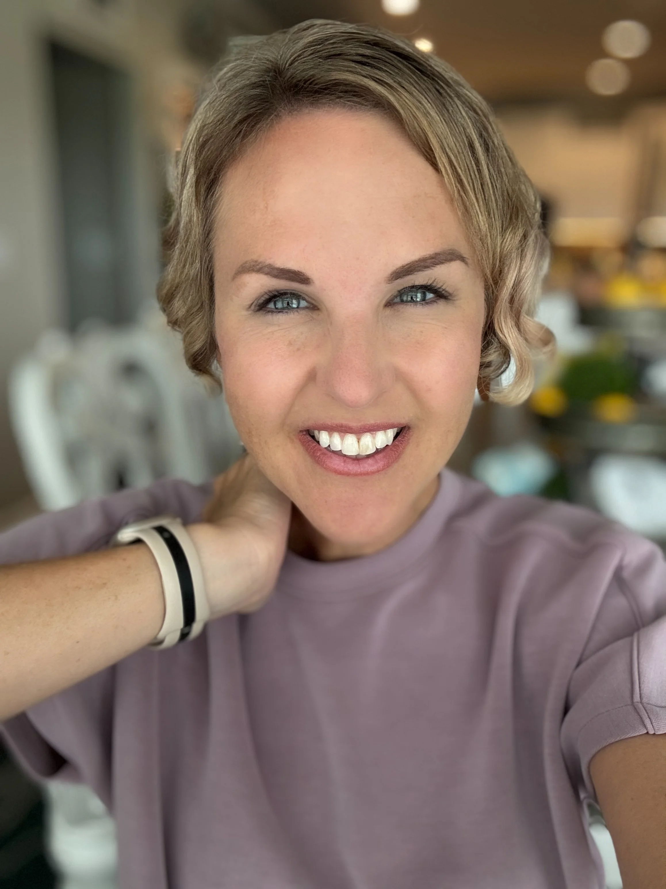 A woman with short blonde hair and blue eyes smiling, wearing a mauve shirt and a fitness tracker on her wrist, in a bright indoor setting.