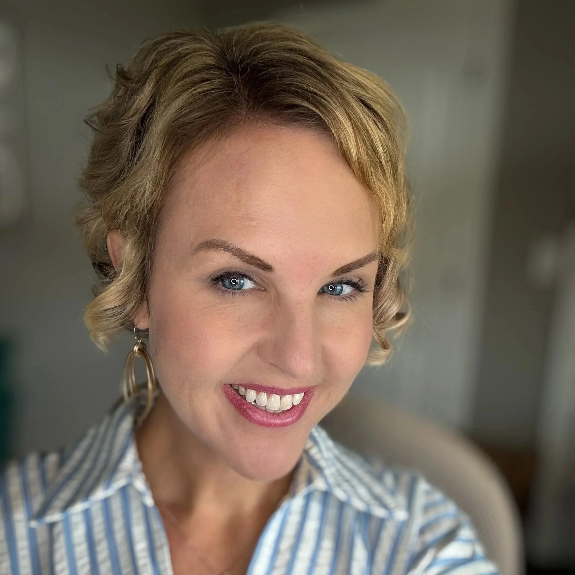 Close-up of a smiling woman with blue eyes, short wavy blonde hair, wearing a striped collared shirt and gold hoop earrings.