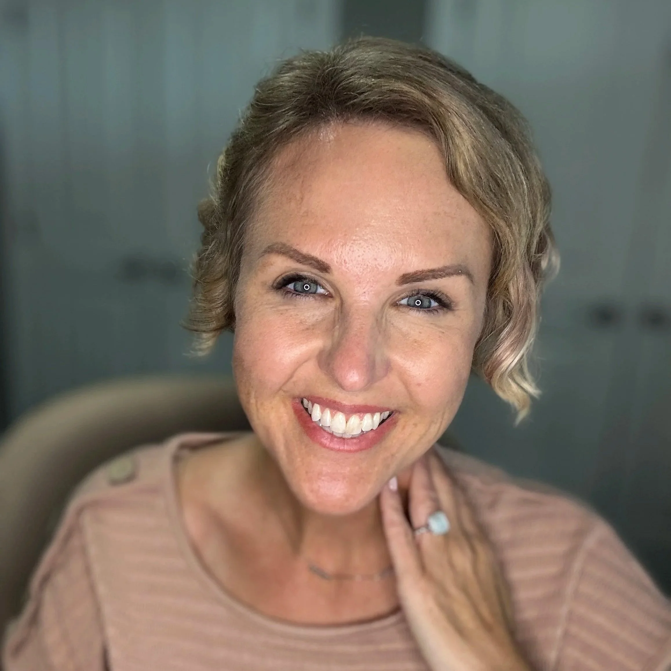 A woman with short wavy blonde hair, blue eyes, and light skin smiling at the camera in an indoor setting.