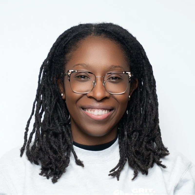 A woman with dark skin, glasses, and dreadlocks smiling at the camera against a white background.