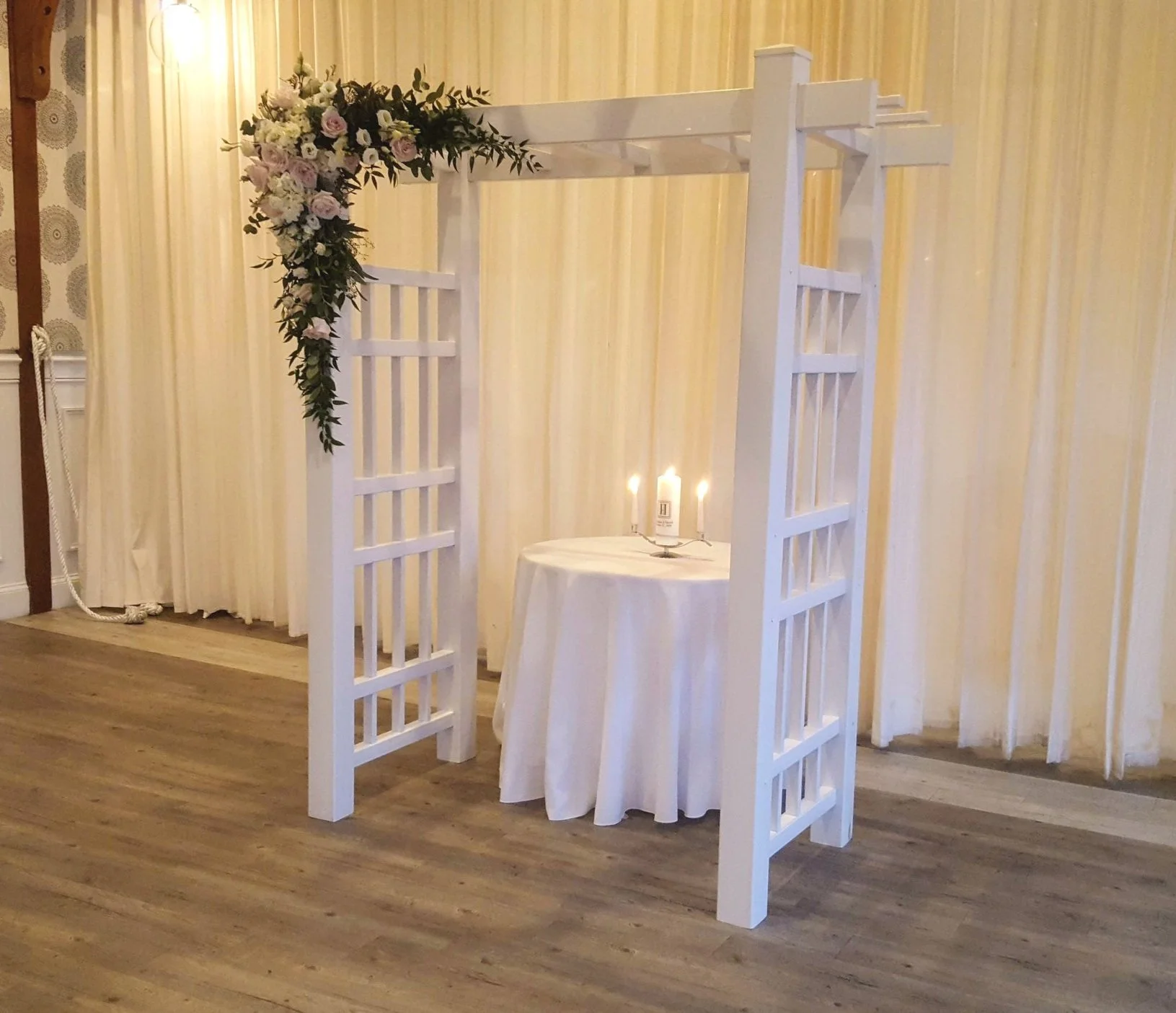 Wedding altar with a white wooden arch decorated with pink and white flowers and greenery, a small round table with candles, and a yellow striped curtain in the background.