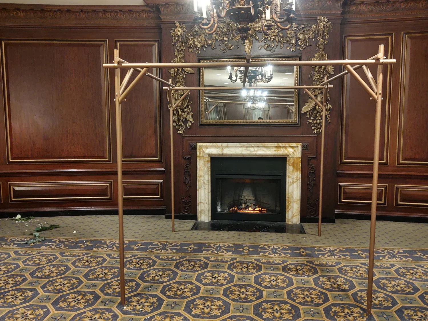 Indoor scene with a decorative wooden wall, a marble fireplace, an ornate mirror, and a chandelier. A metal frame is positioned in front of the fireplace, and the floor has a patterned carpet with some scattered leaves or debris.