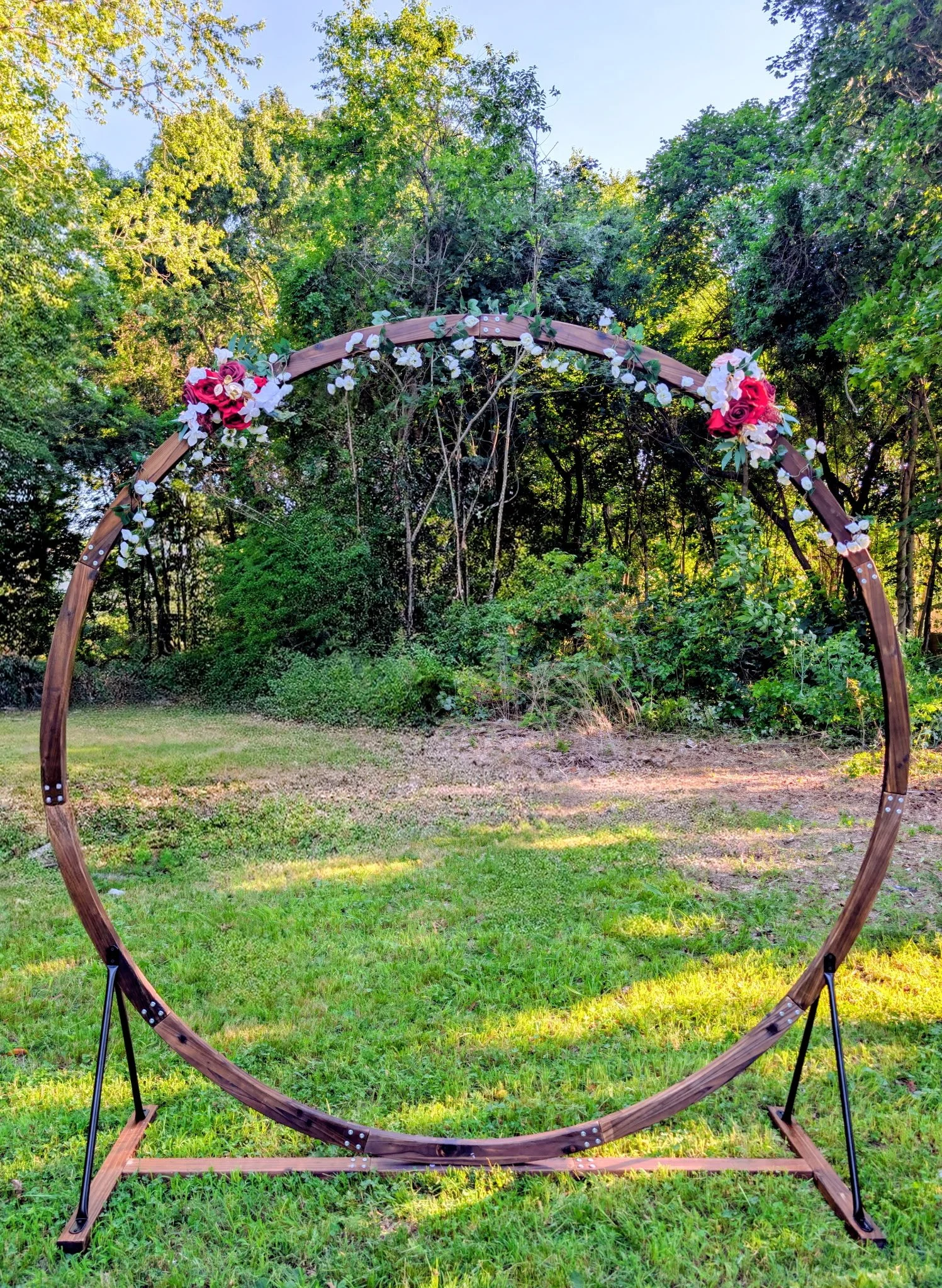 A wooden arch decorated with pink and white flowers set up outdoors on a grassy area, surrounded by trees and greenery, with sunlight filtering through the leaves.