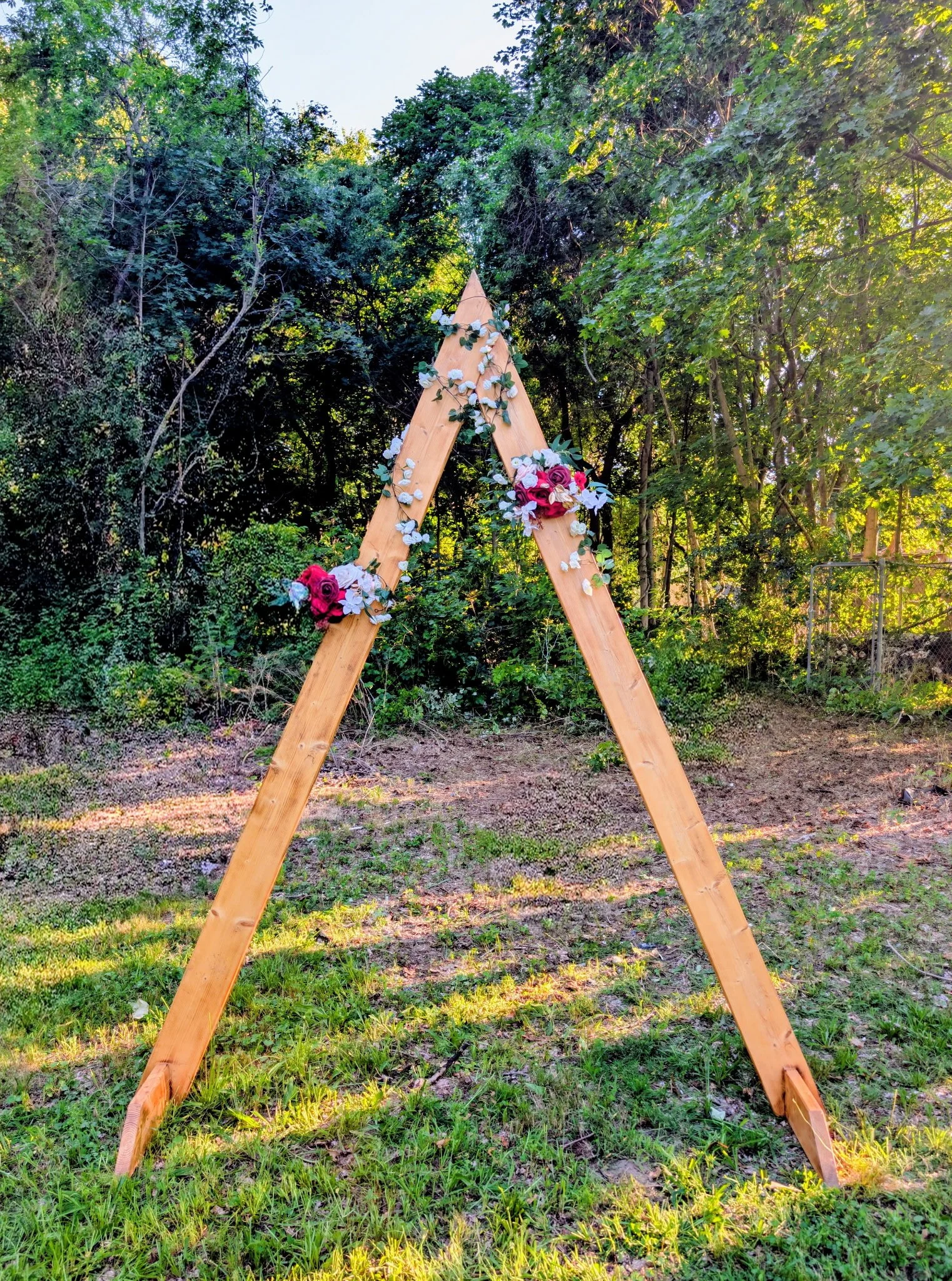 A wooden wedding arch decorated with white, pink, and purple flowers stands in a lush green outdoor setting surrounded by trees and grass.