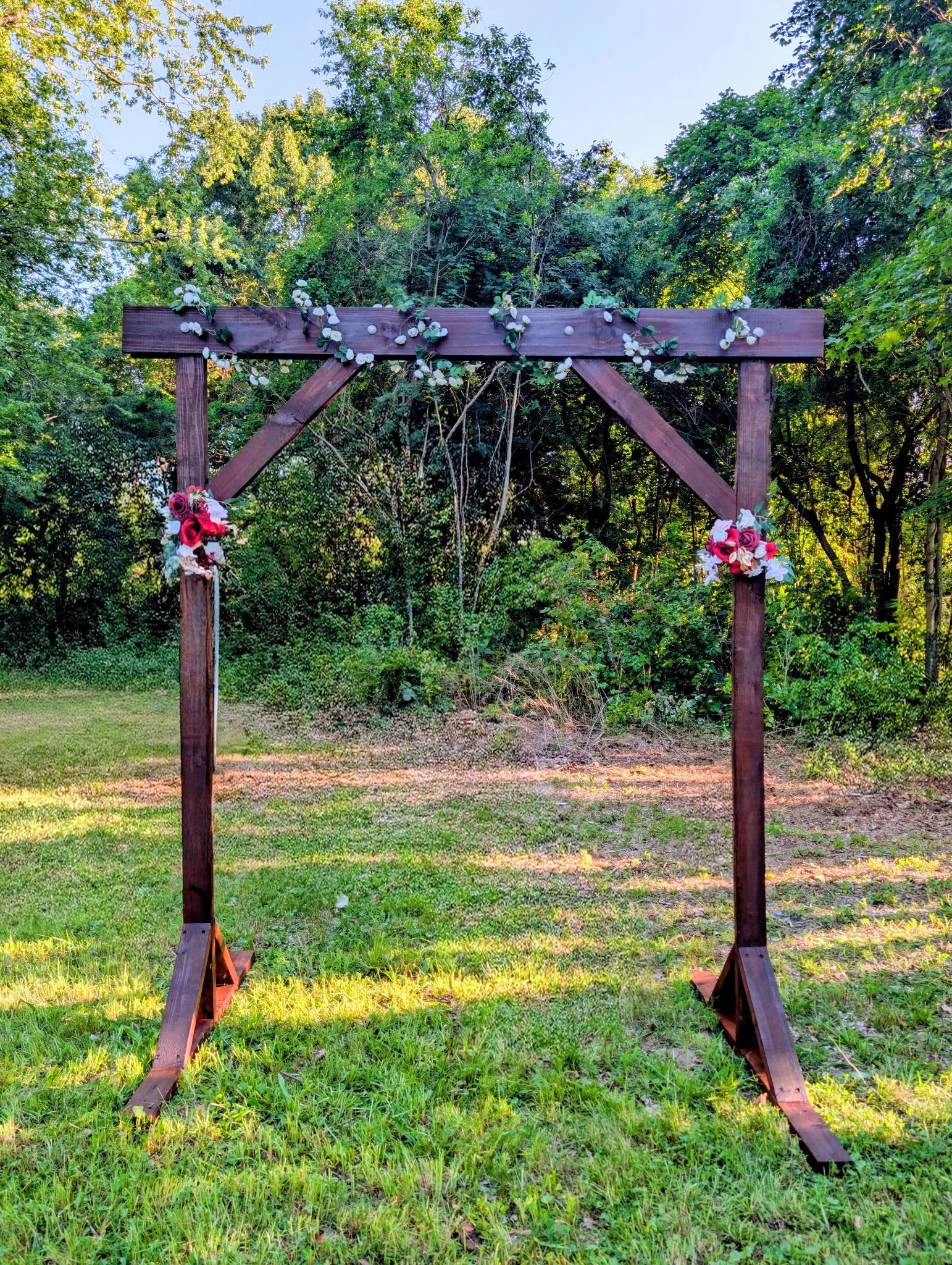A wooden arch decorated with pink and white artificial flowers set in a green outdoor area with trees and grass.