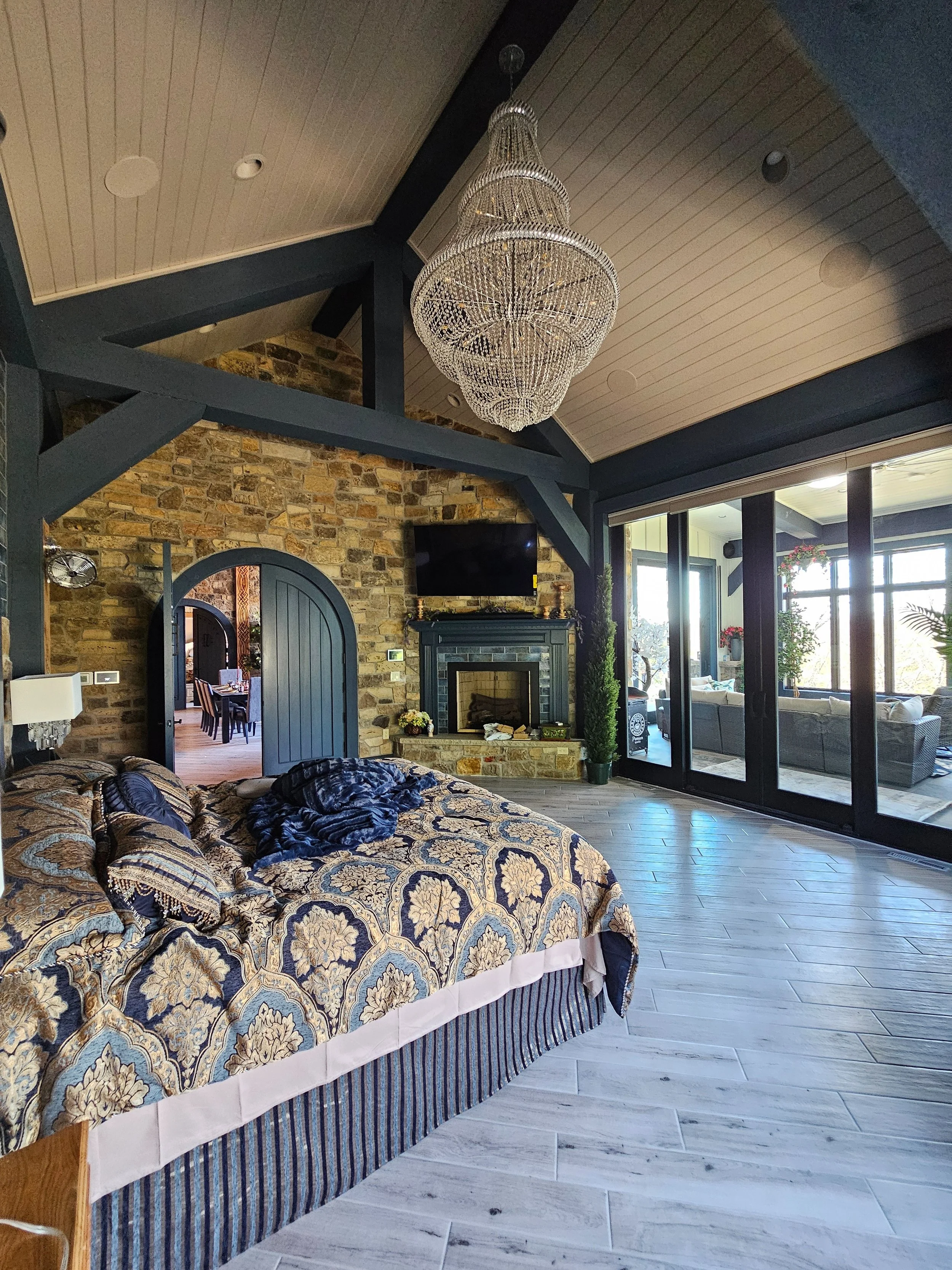 A spacious bedroom with a large bed covered in ornate bedding, a chandelier hanging from the vaulted ceiling, a stone fireplace with a TV mounted above, and sliding glass doors leading to an outdoor patio with seating.