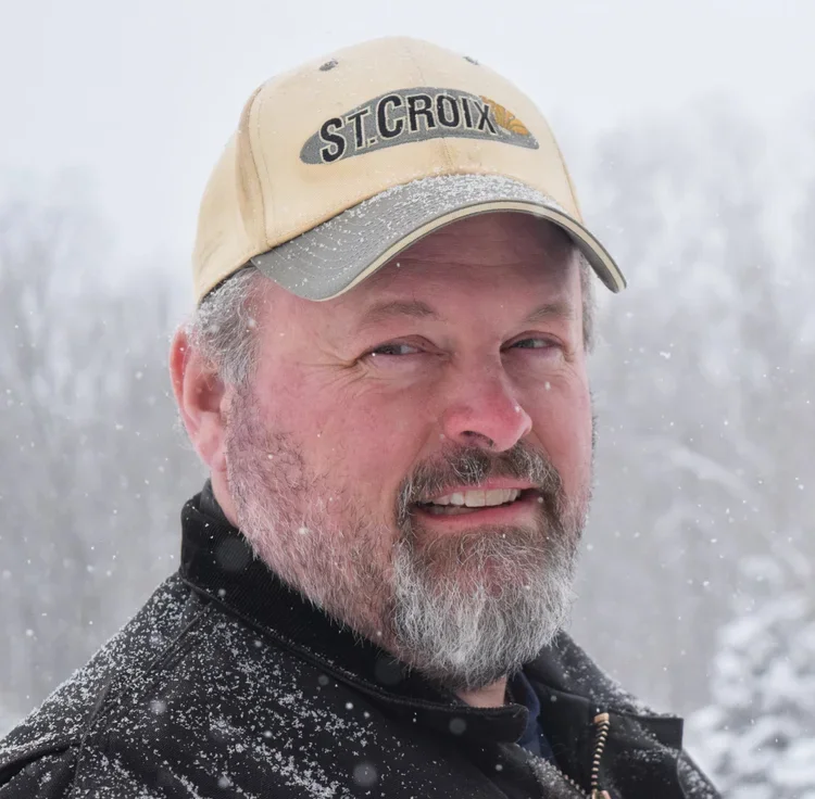 A smiling man with a beard in snowy weather, wearing a beige cap and black jacket.