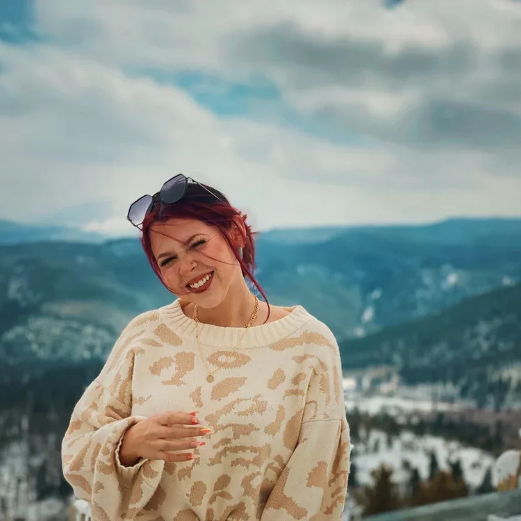 A woman with red hair, wearing a cream and beige patterned sweater, sunglasses on her head, and smiling outdoors with mountains and a cloudy sky in the background.