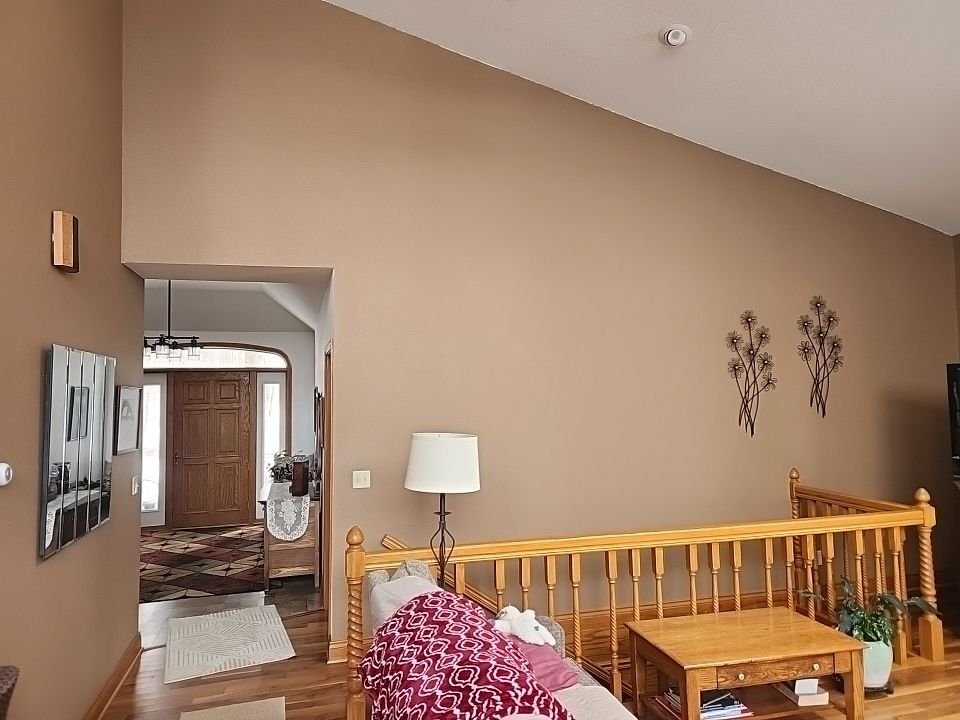 Living room with beige walls, decorated with metal wall art, wooden furniture including a side table and railing, a lamp, a pink and white patterned blanket on a sofa, and an entryway with a wooden door and a rug.