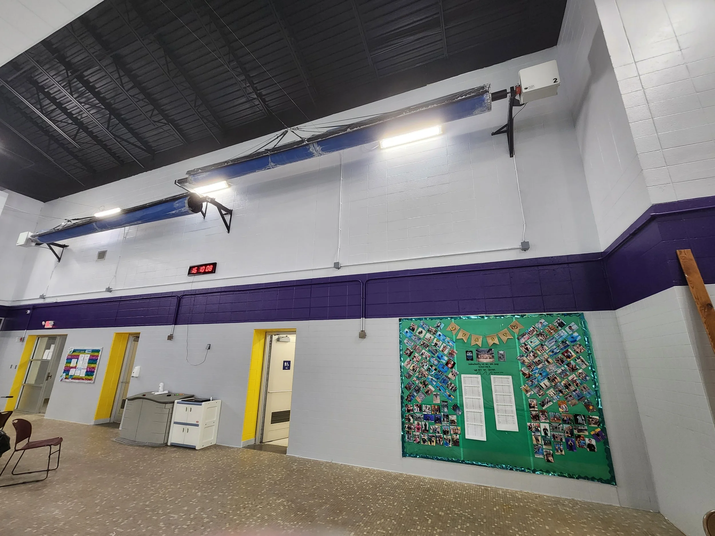 Interior of a gymnasium or community center with white and purple walls, a black ceiling, and various bulletin boards. There are two doors, a water fountain, chairs, and a display board with photos.