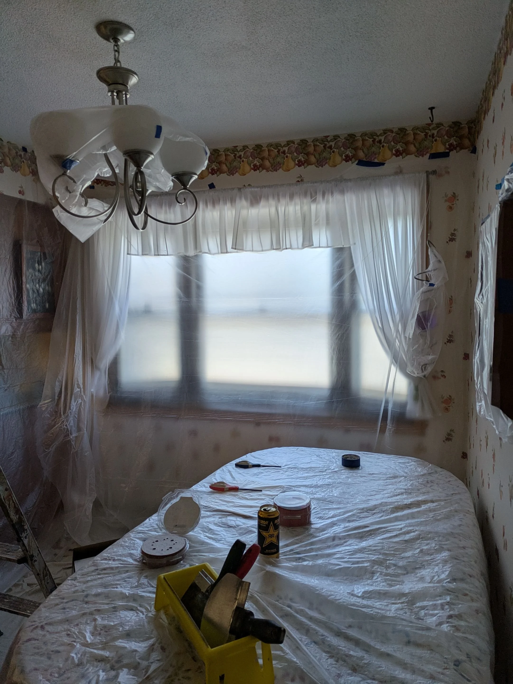 Room undergoing renovation, with plastic covering furniture and window, tools and supplies on the table, and a chandelier hanging from the ceiling.