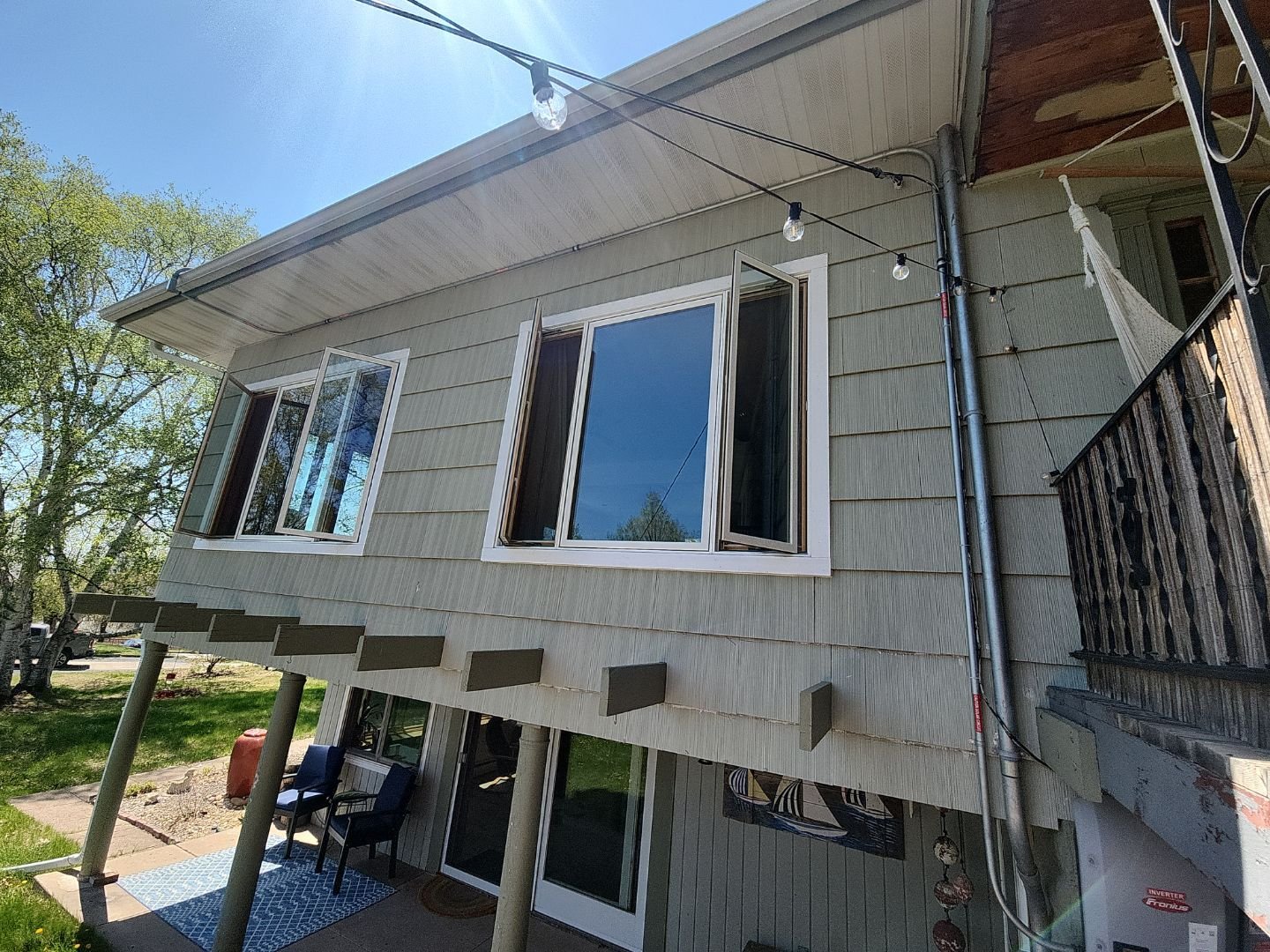 The image shows the exterior of a house with beige siding and a second-story balcony. The balcony has a black railing, and there is a hammock hanging on it. Two large open windows reveal a clear blue sky and trees outside. Strings of outdoor string lights are hung across the balcony area, and a drainpipe runs vertically down the house's corner. Below, there is a patio with chairs and a small mat, with trees and grassy area in the background.