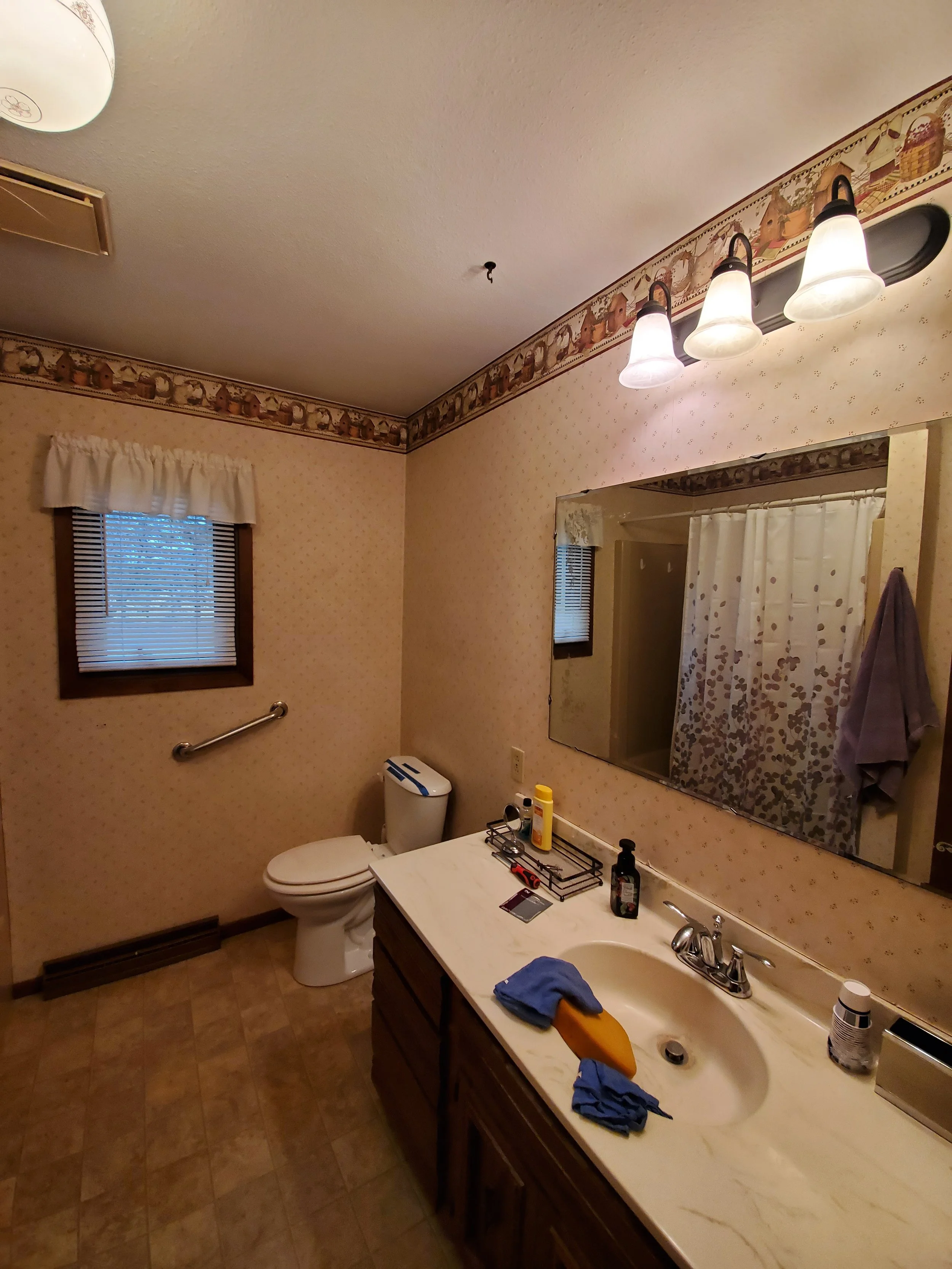 A bathroom with beige wallpaper, a small window with a white curtain, a large mirror, a light fixture with three bulbs, a white toilet, and a wooden vanity with a marble top. There are cleaning cloths on the sink, and various toiletries and supplies on the counter.