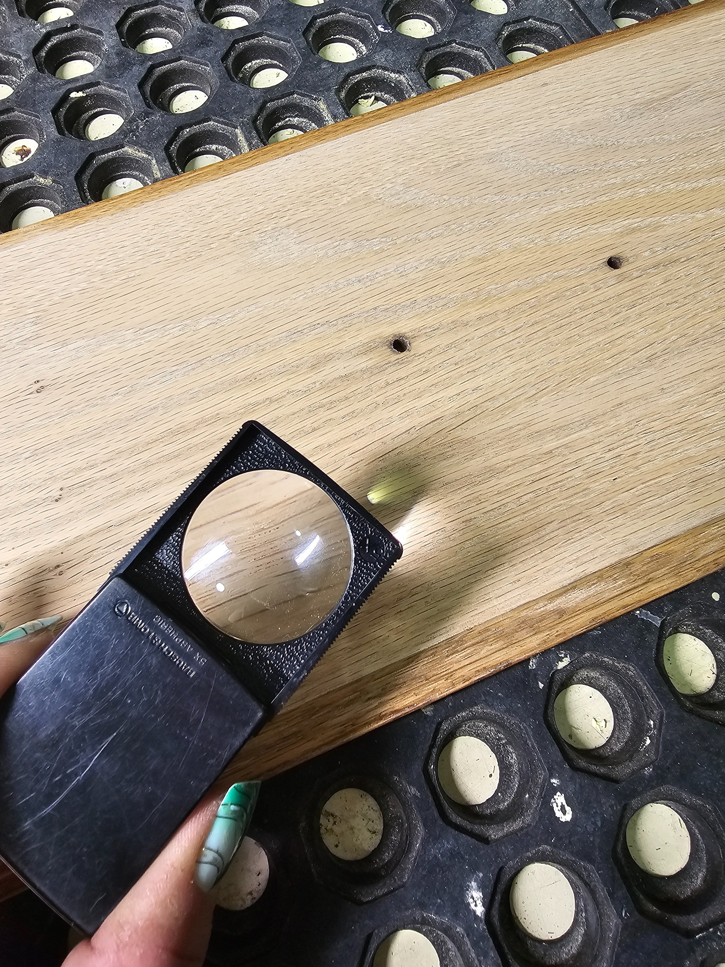 A hand holding a magnifying glass over a wooden surface with a small hole, with a rubber mat with white circular objects in the background.
