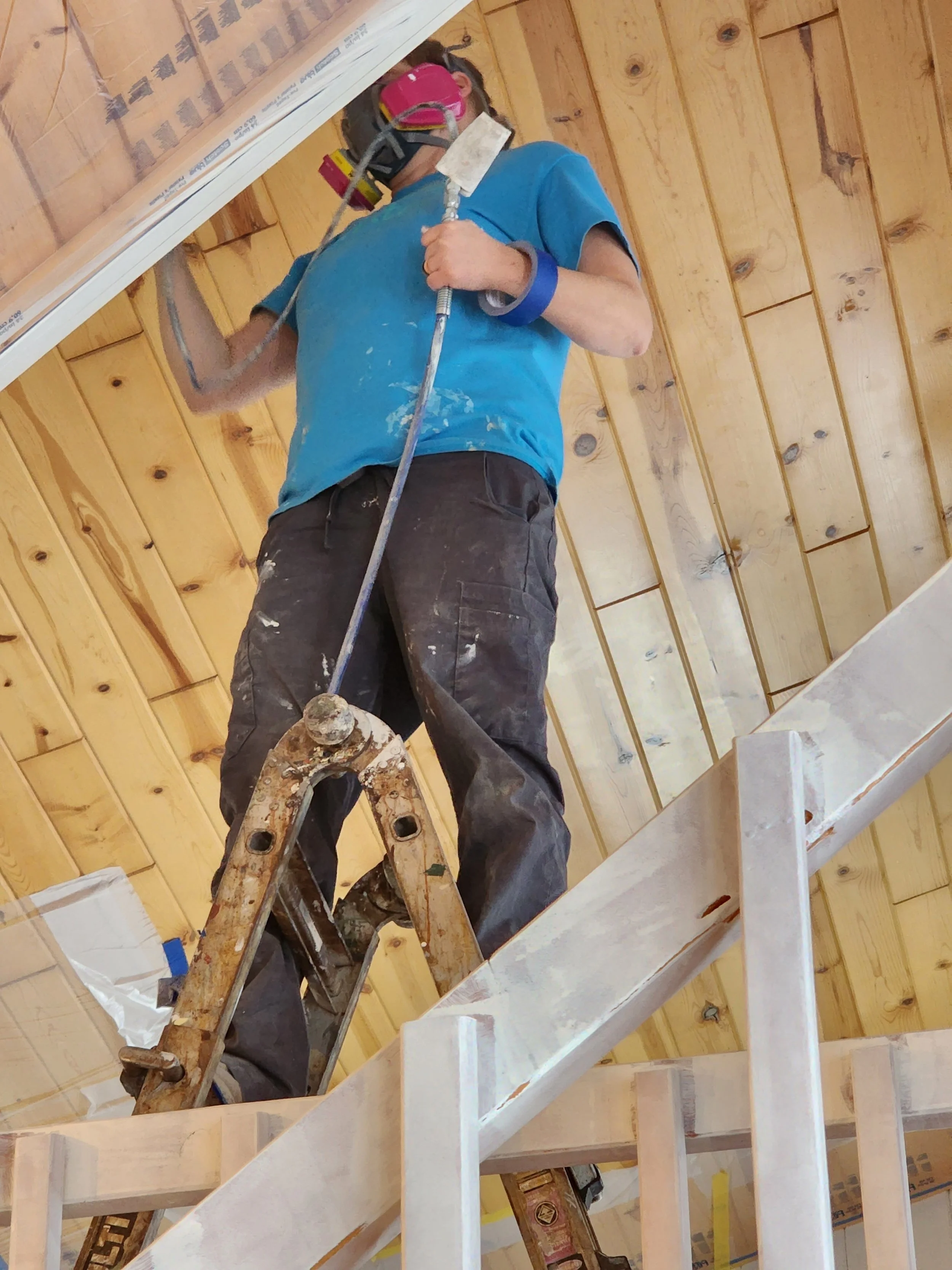 Person wearing a blue shirt, pink safety goggles, and a respirator mask, standing on a wooden ladder while sanding or painting a ceiling with a paint roller or brush.