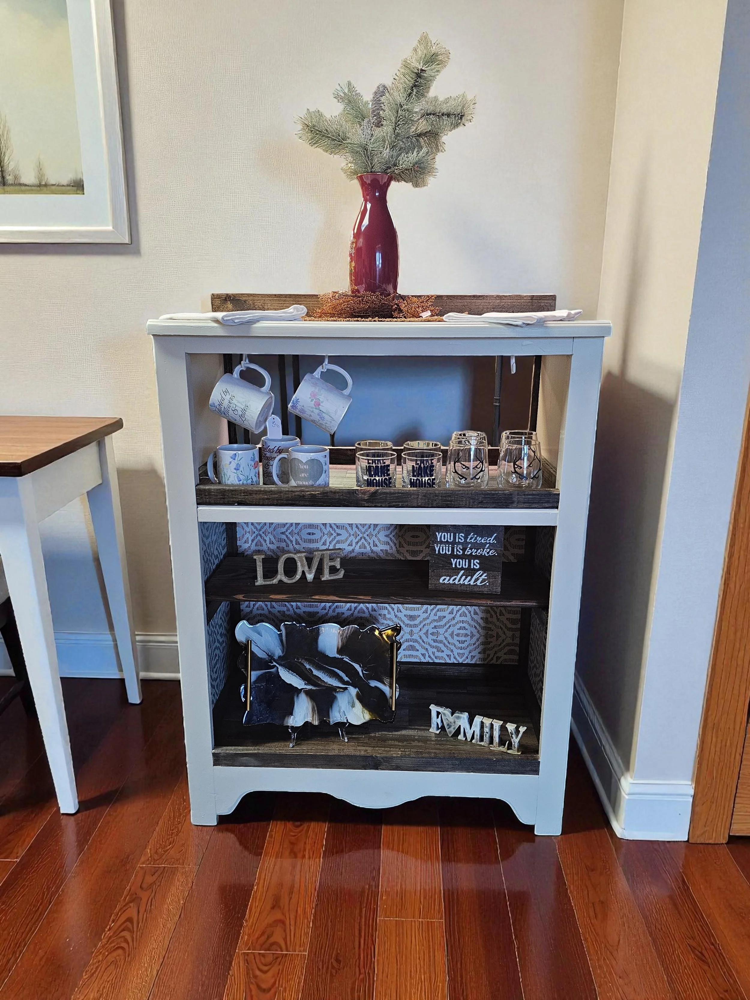 A decorative white and dark wood shelf unit with various mugs, glasses, and decorative items, topped with a red vase holding pine branches, against a beige wall with a framed picture.