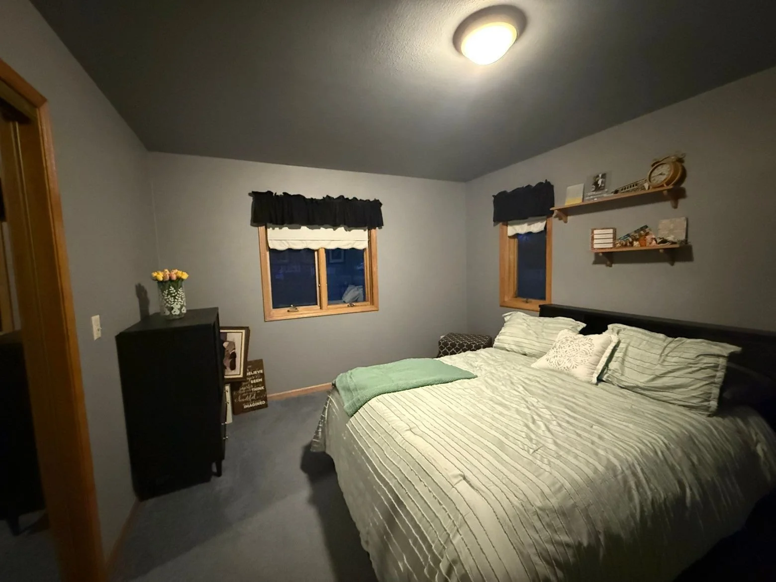 A bedroom with blue walls, a bed with striped bedding, two windows with black valances, a black dresser with a vase of yellow flowers, and wall shelves holding various items.
