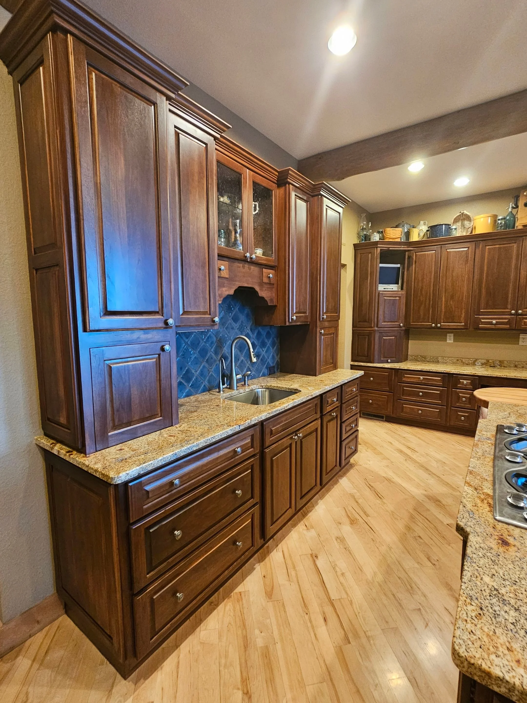 Kitchen with dark wood cabinetry, granite countertops, a small sink, and a blue tiled backsplash. Recessed ceiling lights illuminate the area, and there's a view of additional cabinets and countertop space in the background.