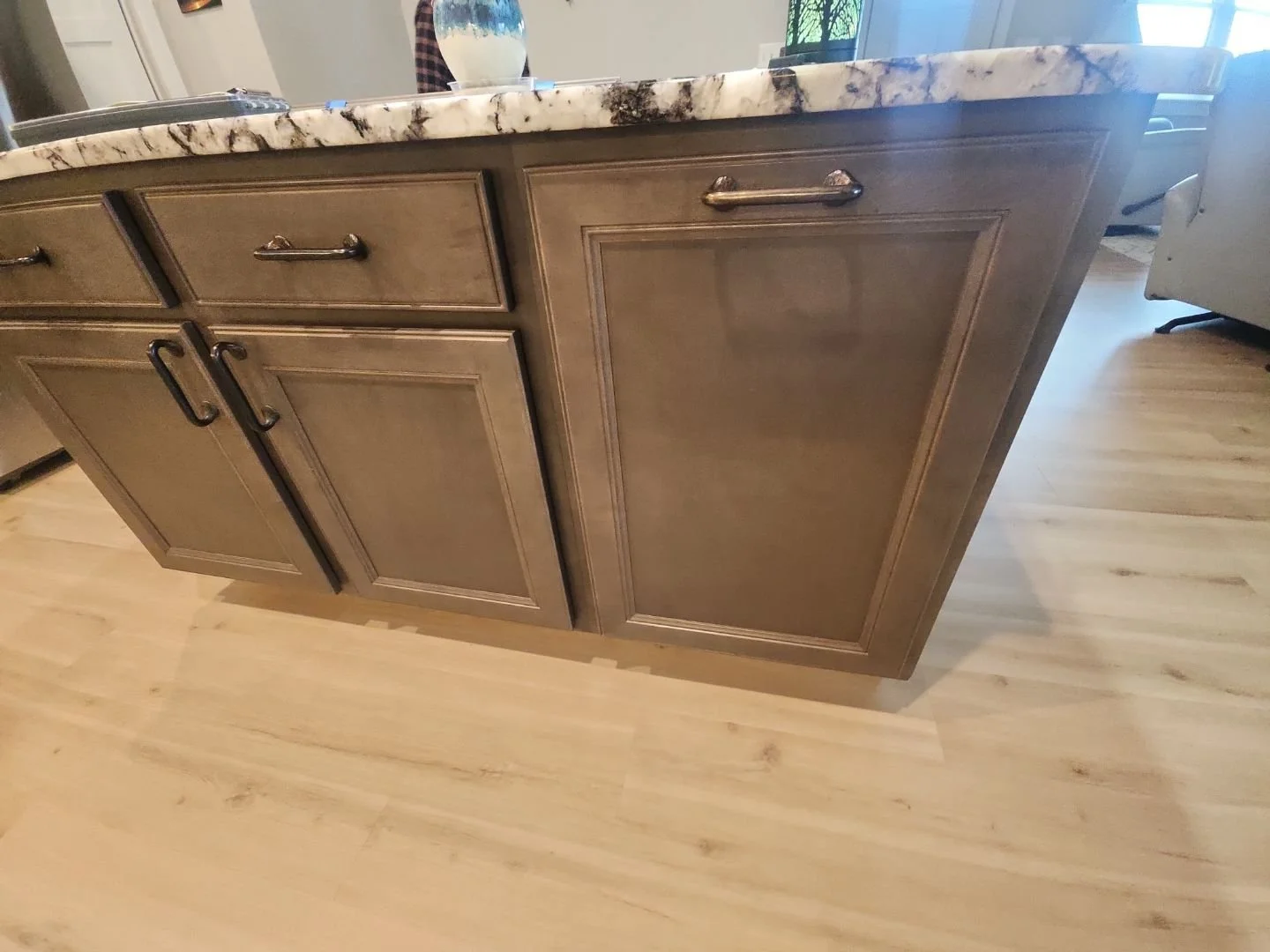 A kitchen island with a marble countertop and beige wooden cabinets with black handles, in a room with light wooden flooring.