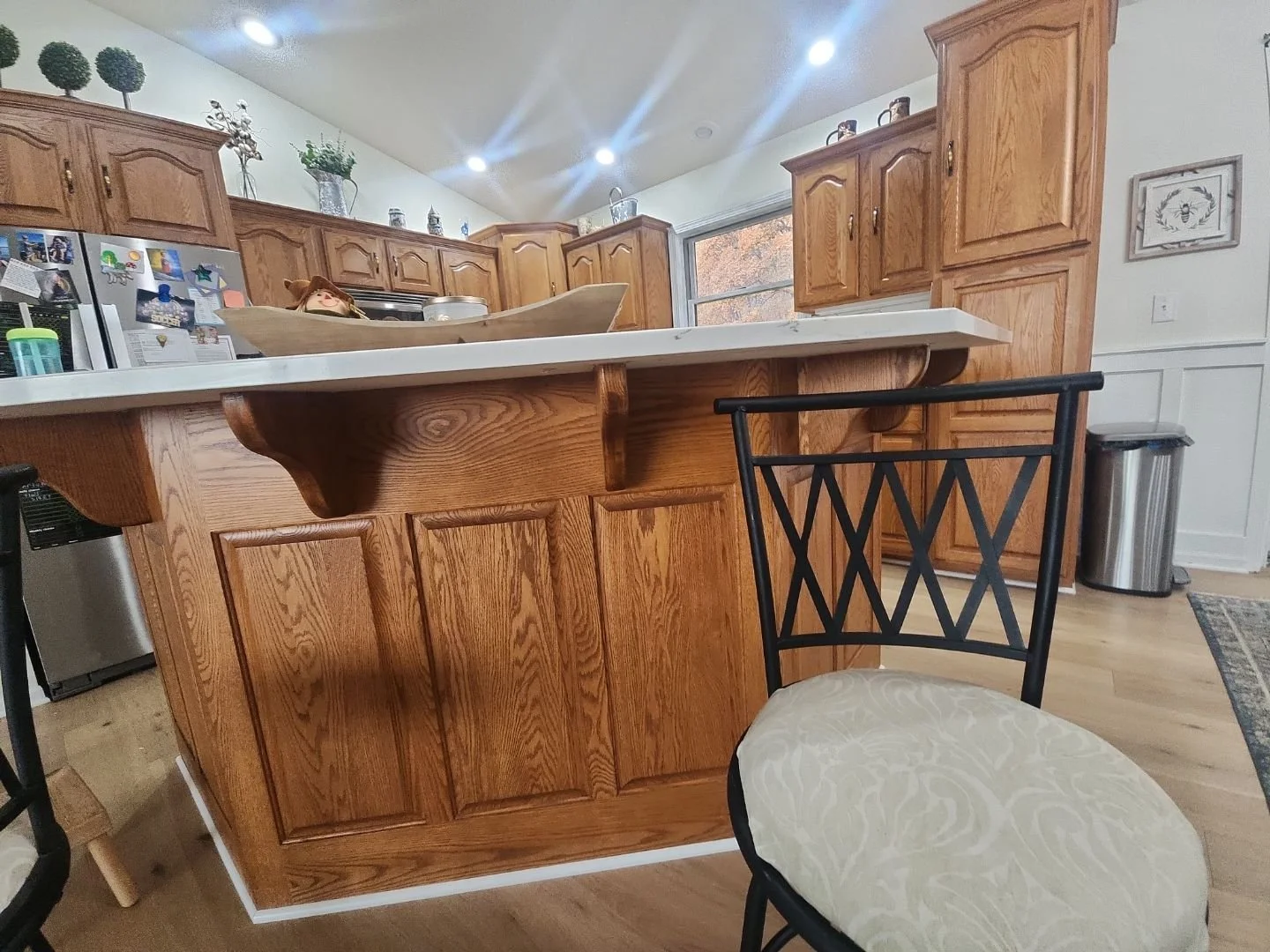 Kitchen with wooden cabinets, a white countertop on a kitchen island, a black chair with an upholstered seat, a stainless-steel trash can, and a window showing autumn trees outside.