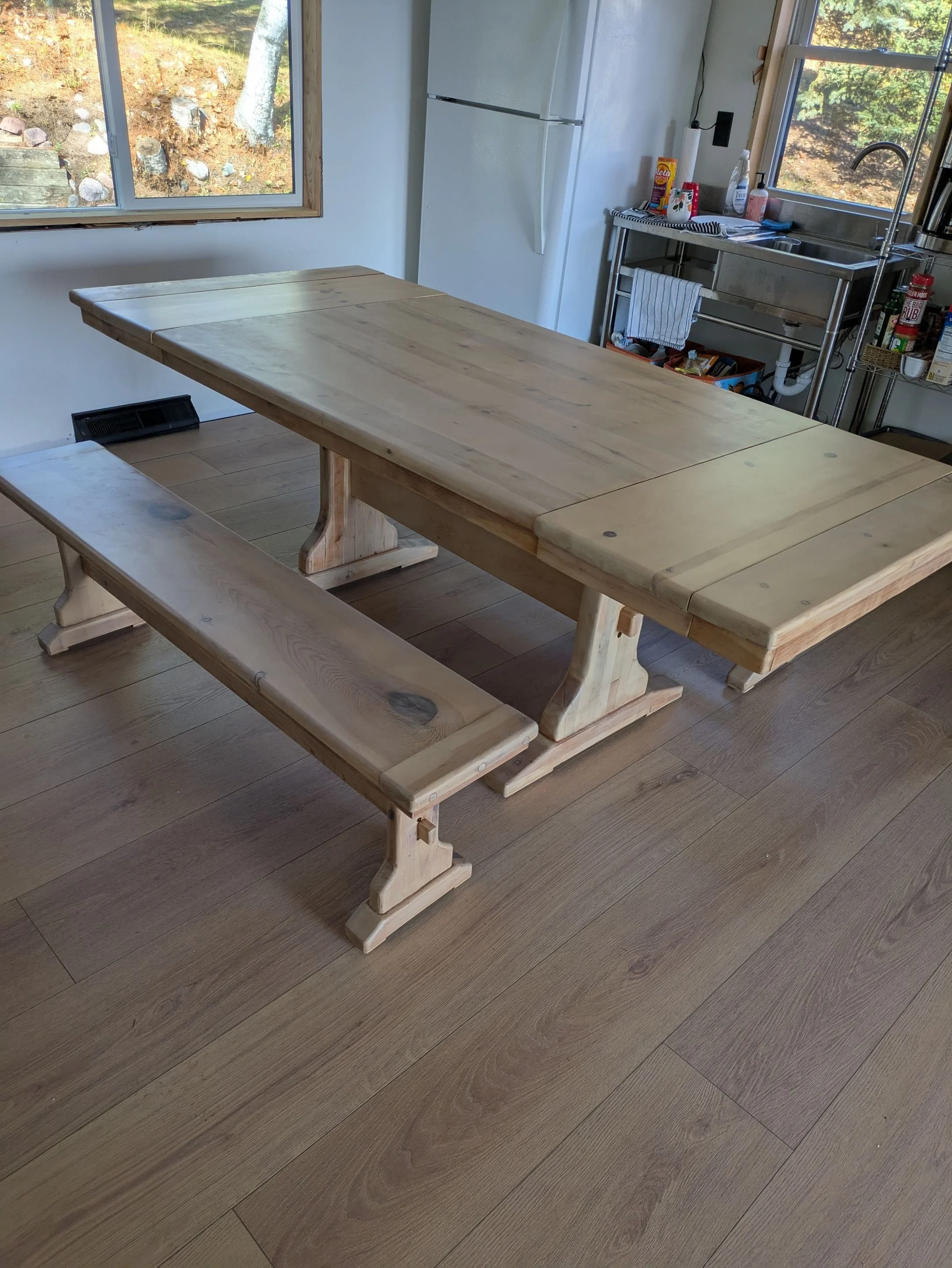A wooden dining table with matching benches in a kitchen with wooden flooring and a window showing trees outside.