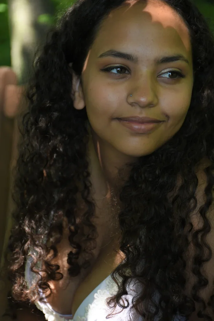 A woman with long, curly black hair and a nose piercing smiling outdoors with blurred greenery in the background.