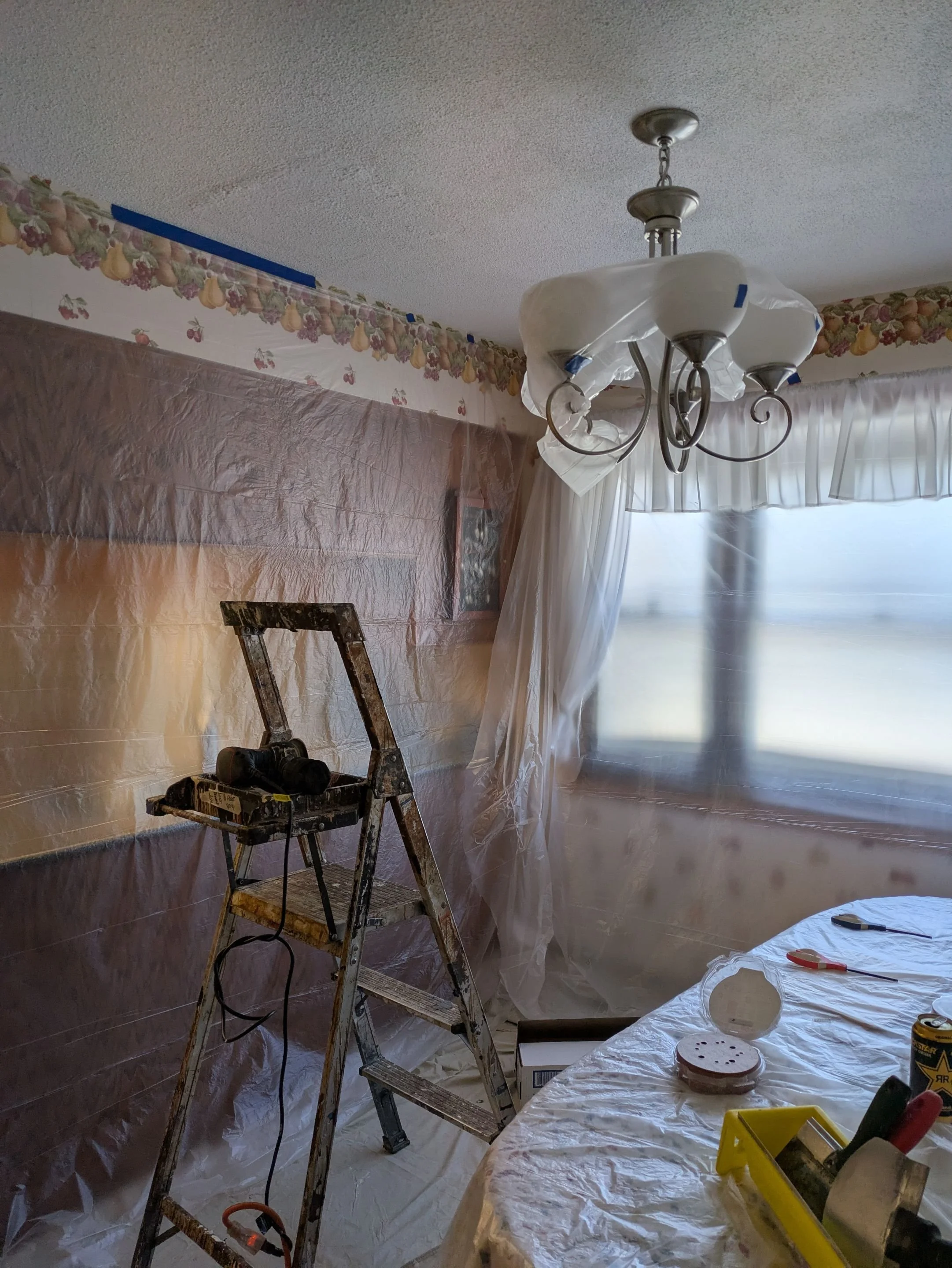 A room under renovation with plastic sheeting covering the walls and a window, a metal step ladder with tools on top, a table with tools and supplies, and a ceiling light fixture.