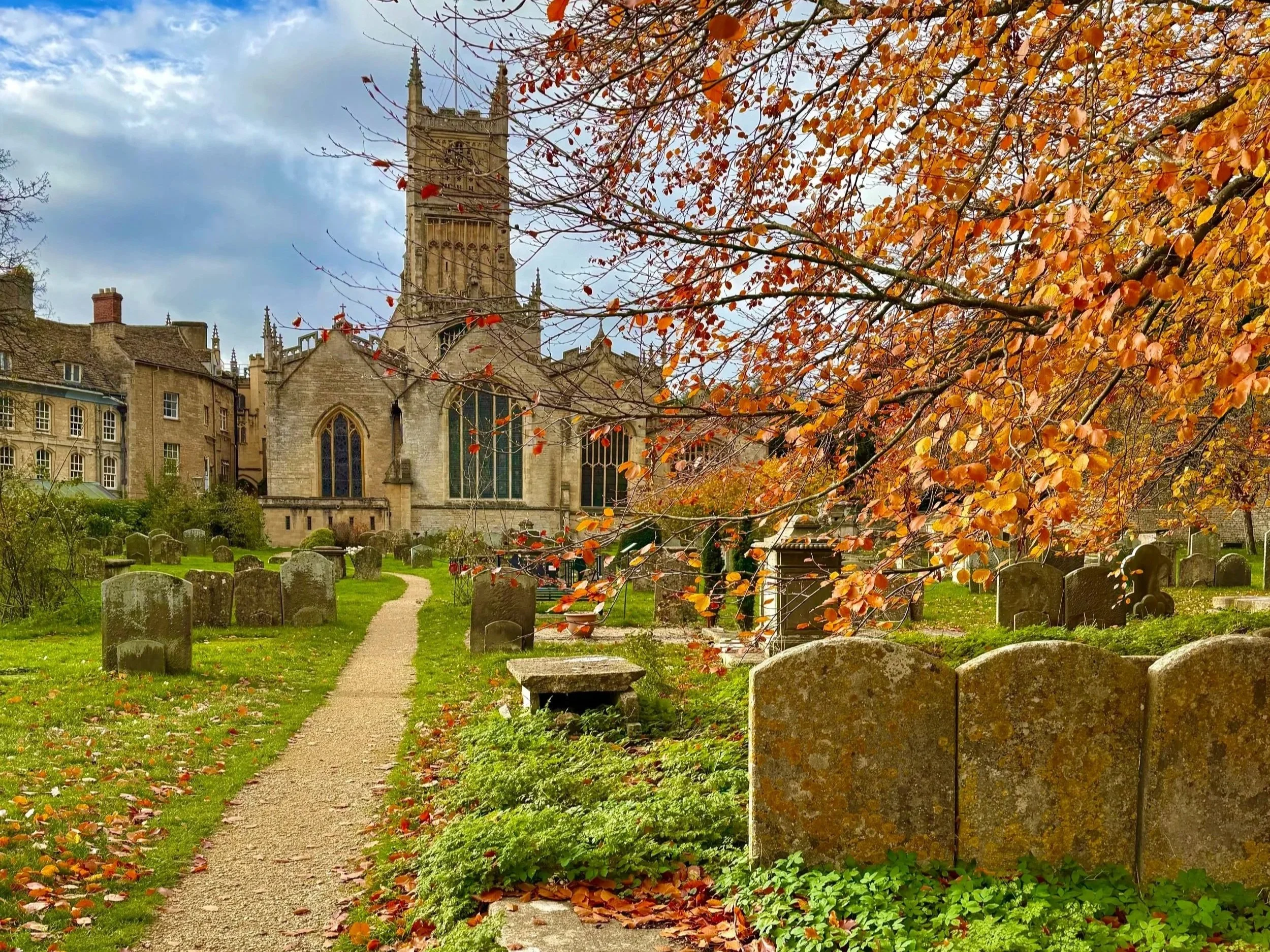 Autumn scene of a churchyard with tombstones, a pathway, and a large tree with orange leaves in front of a historic church.