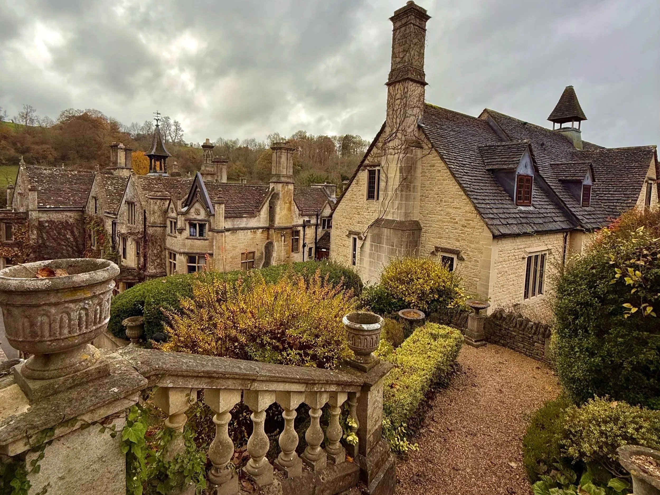 Charming stone houses with slate roofs, surrounded by lush bushes and trees, viewed from a stone balcony with ornate balusters in Castle Combe UK