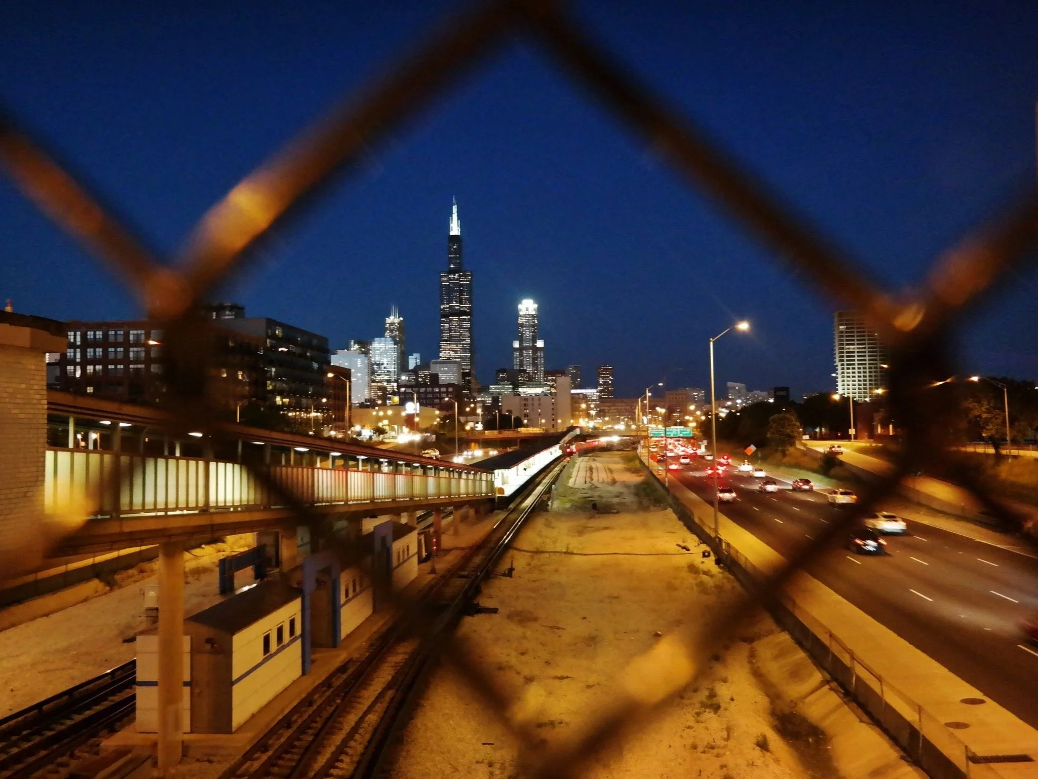 Night view of the Chicago skyline through a chain-link fence, featuring the Willis Tower and city lights along a busy highway.