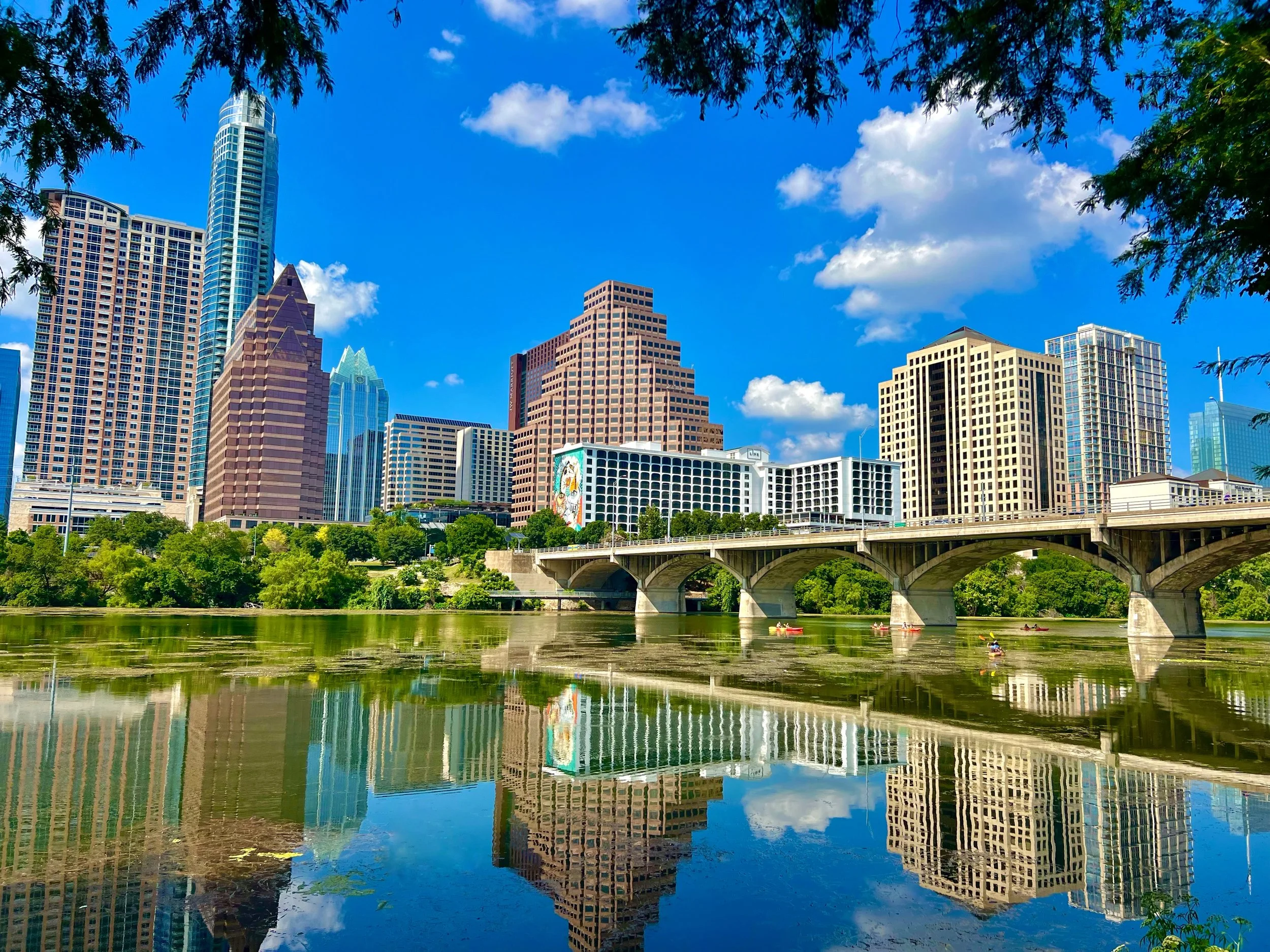 Downtown Austin skyline with skyscrapers, a bridge over a river, and boats with kayakers under a blue sky with clouds.