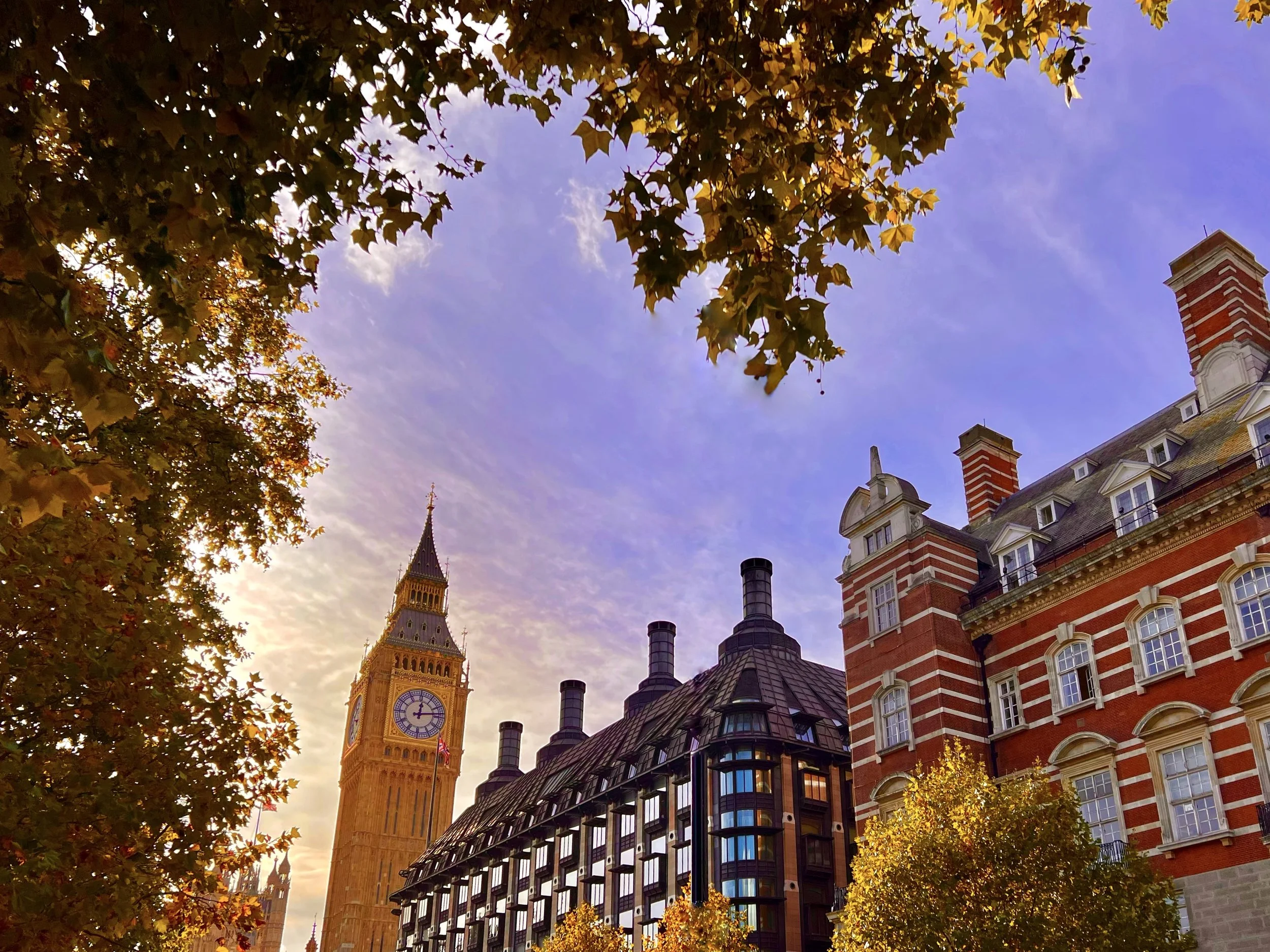 Big Ben clock tower in London with autumn trees in the foreground and a partly cloudy sky.