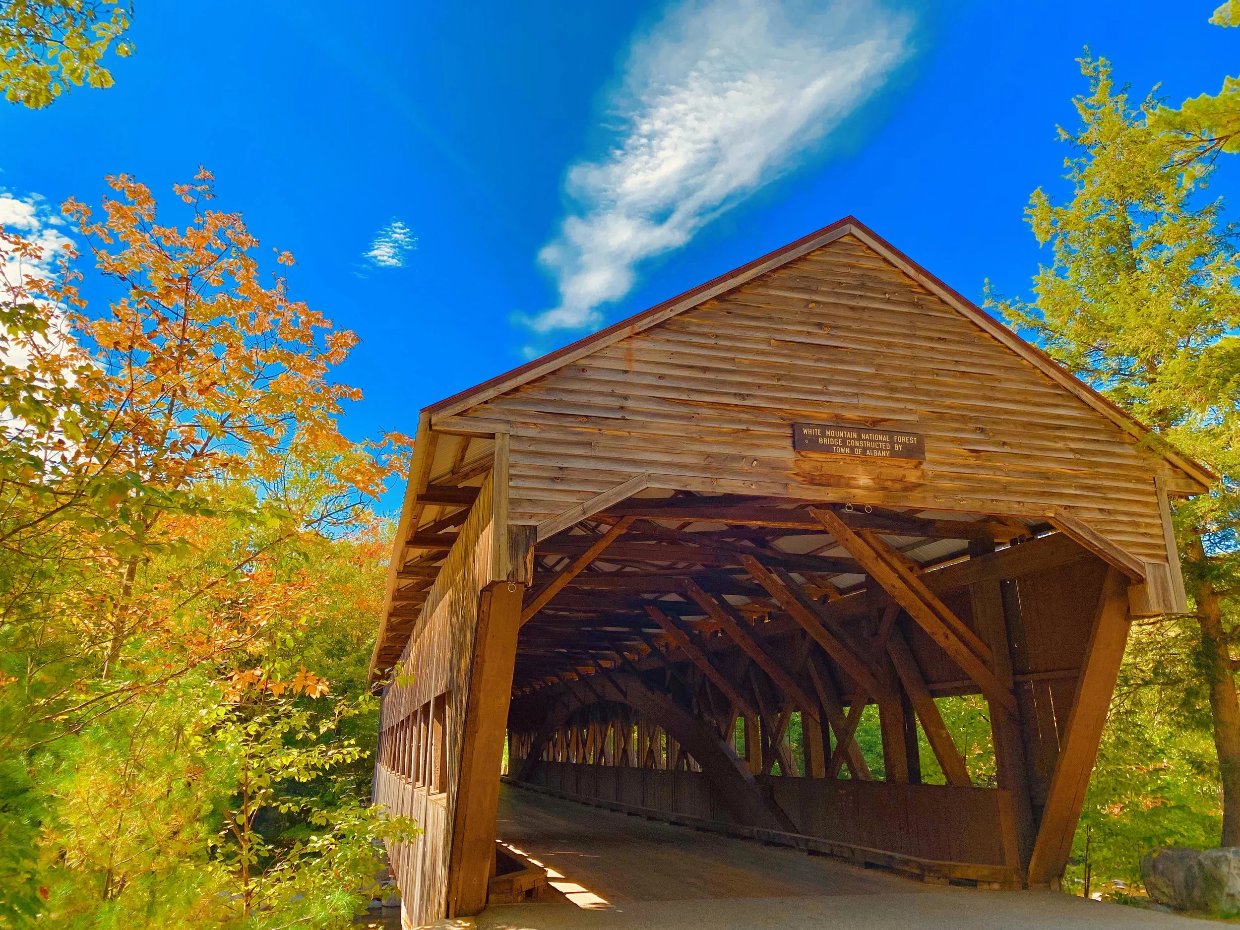 A wooden covered bridge surrounded by trees with fall foliage under a blue sky with clouds.