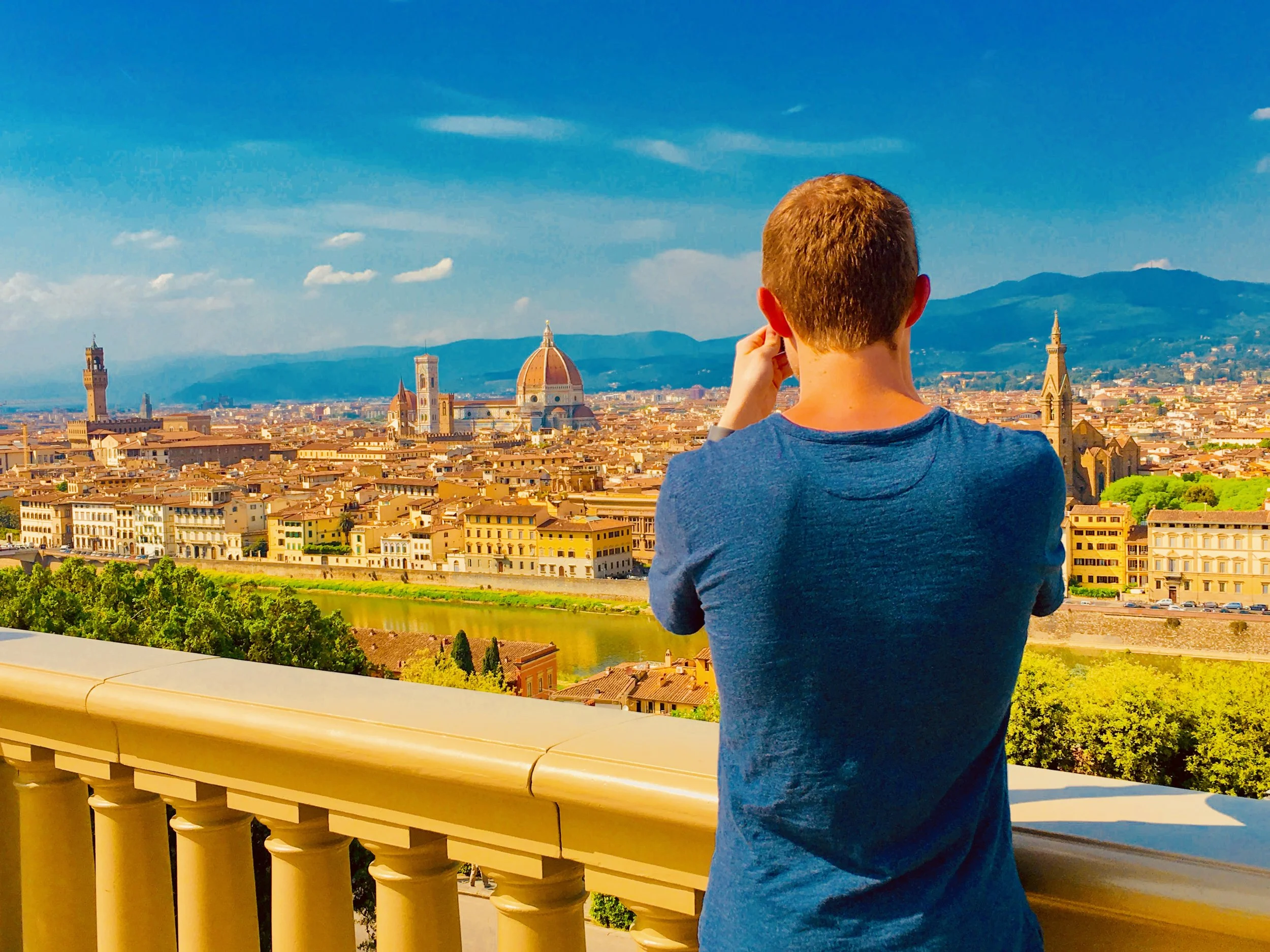 A person with short hair, wearing a blue shirt, standing on a balcony overlooking the city of Florence, Italy. The skyline features the Florence Cathedral with its prominent dome, and the city is situated along a river with mountains in the background.