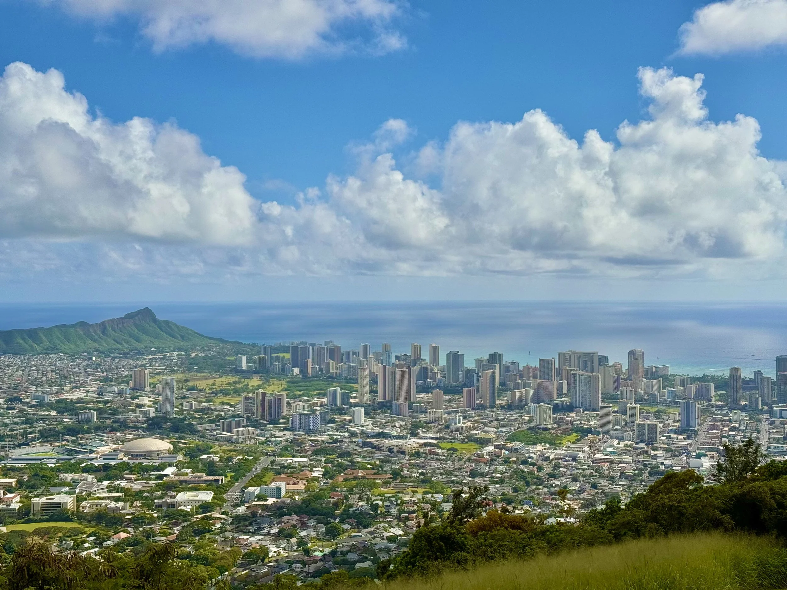 A panoramic view of Honolulu, Hawaii, with high-rise buildings, lush green mountains, and the Pacific Ocean in the background under a partly cloudy sky.