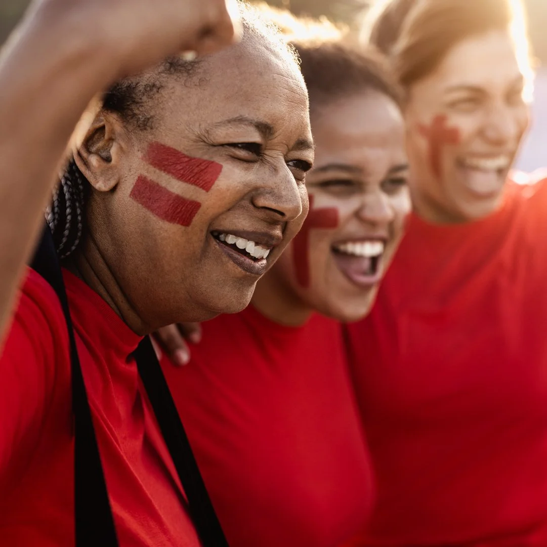Three women with painted red stripes on their cheeks, wearing red shirts, smiling and celebrating together.