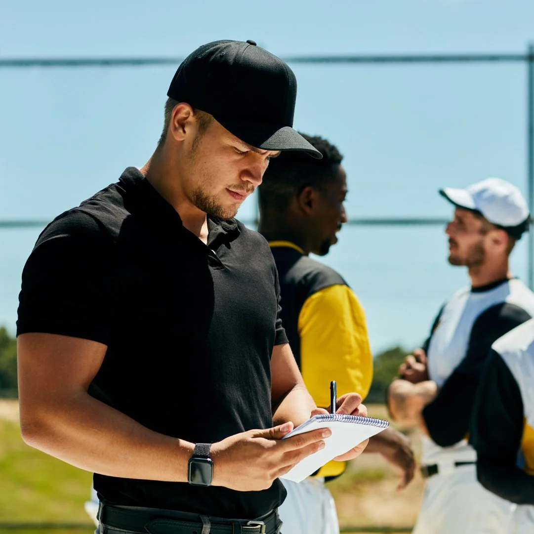 A young man in a black cap and shirt writing in a notebook outdoors, with two baseball players in the background talking to each other.