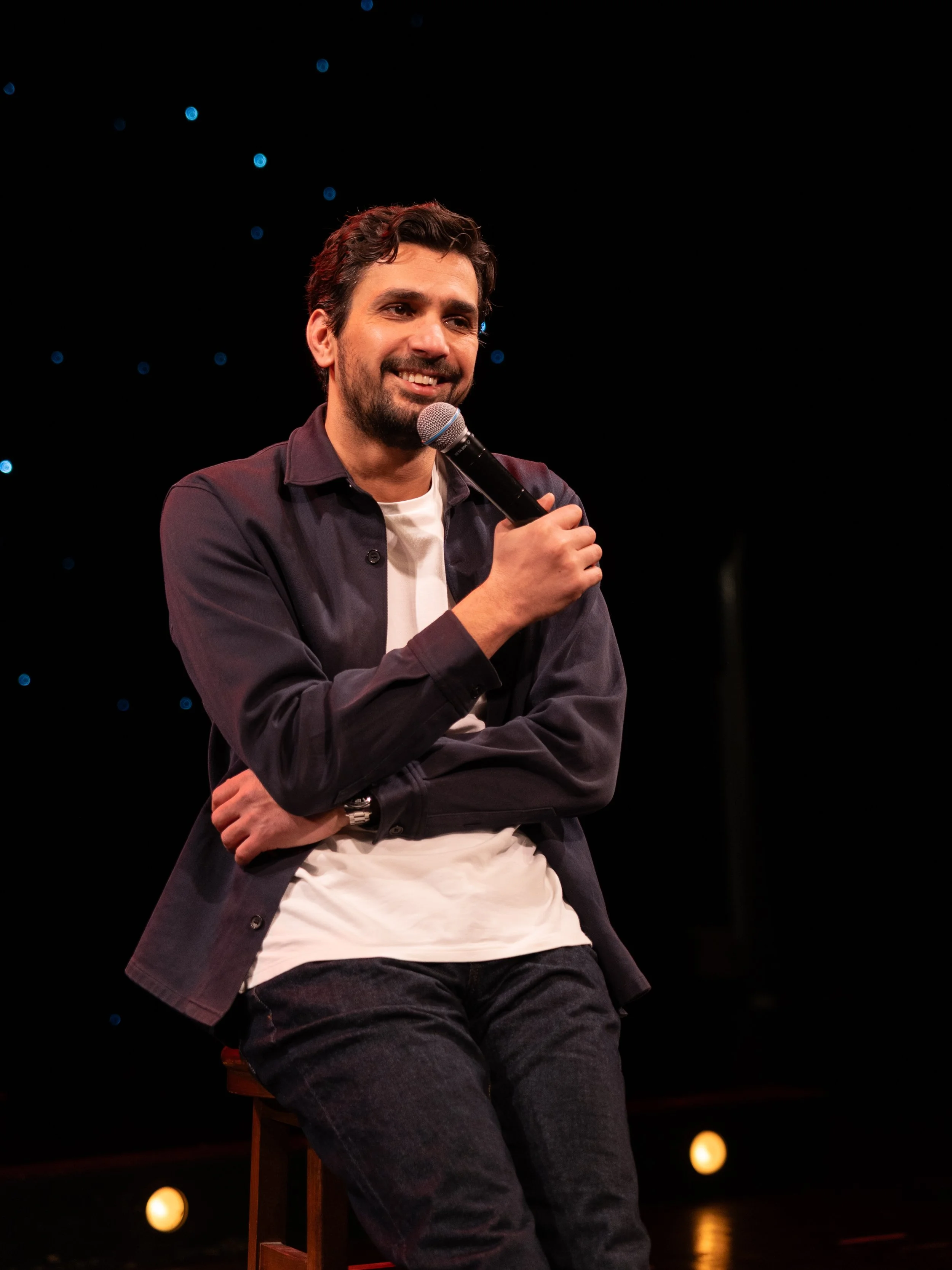 Alaa Abu Diab sitting on a stool, holding a microphone, smiling, against a dark background with small blue lights.