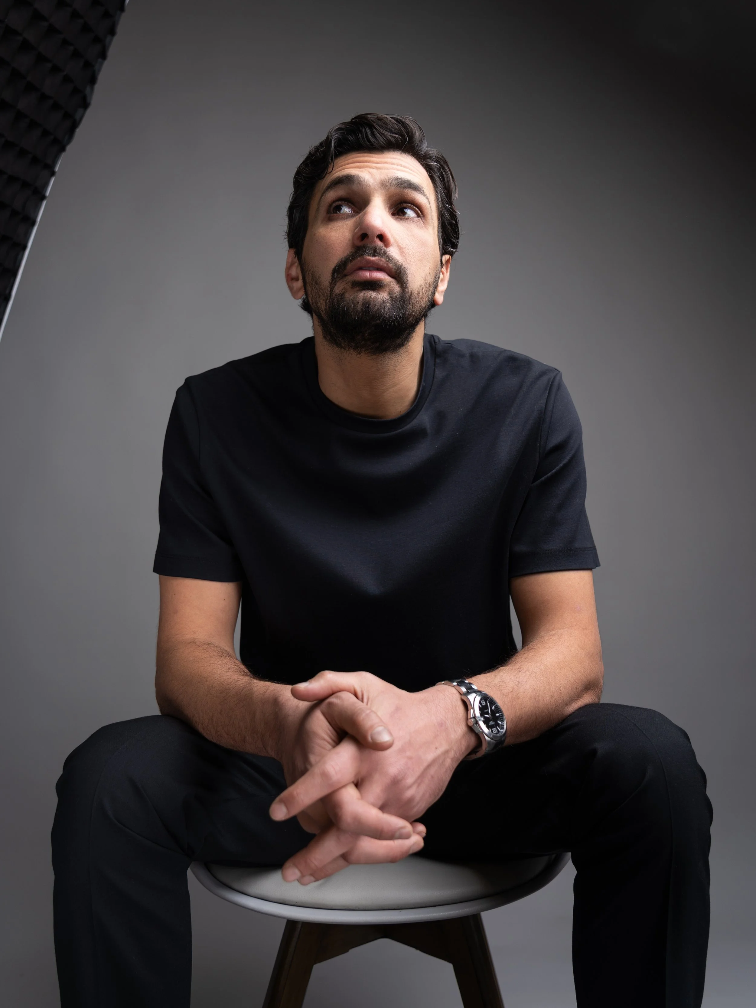 A man with dark hair, beard, and mustache, wearing a black T-shirt, sitting on a white chair, looking upward with a thoughtful expression, in a studio setting with gray background and soft lighting.