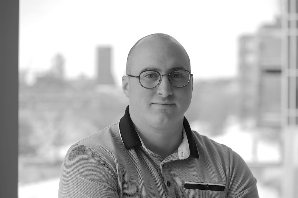 Black and white photo of a man with glasses, wearing a collared shirt, standing indoors with a cityscape in the background.