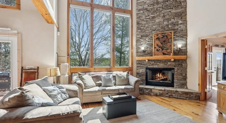 Living room with large window, stone fireplace, beige sofas, wooden floor, and natural light.