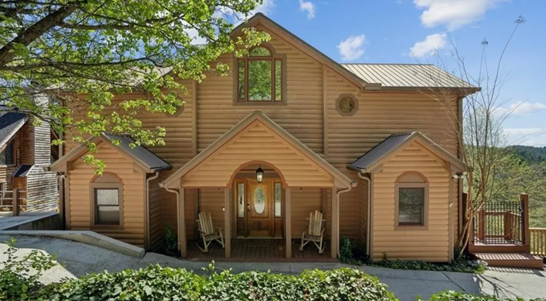 Front view of a two-story wooden house with a covered porch, two wooden chairs, and a decorative front door, surrounded by trees and greenery under a blue sky.