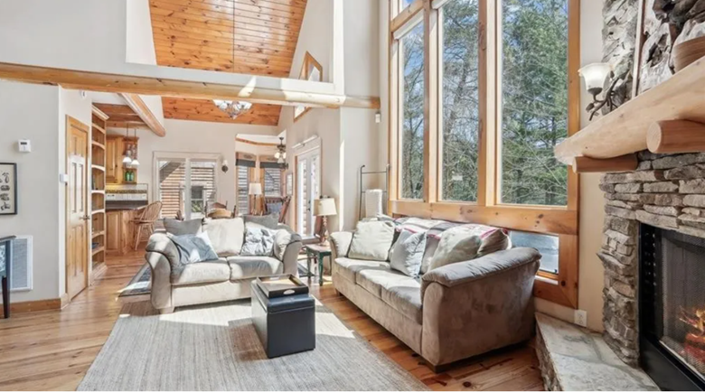 Living room with large windows, stone fireplace, two beige sofas, wooden ceiling, hardwood floors, and a neutral area rug.