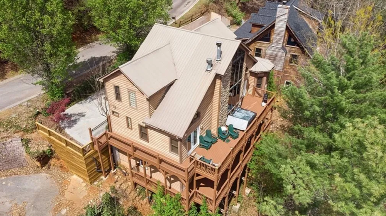 Aerial view of a multi-story wooden house with a large wrap-around deck, surrounded by trees and neighboring houses in a wooded area.